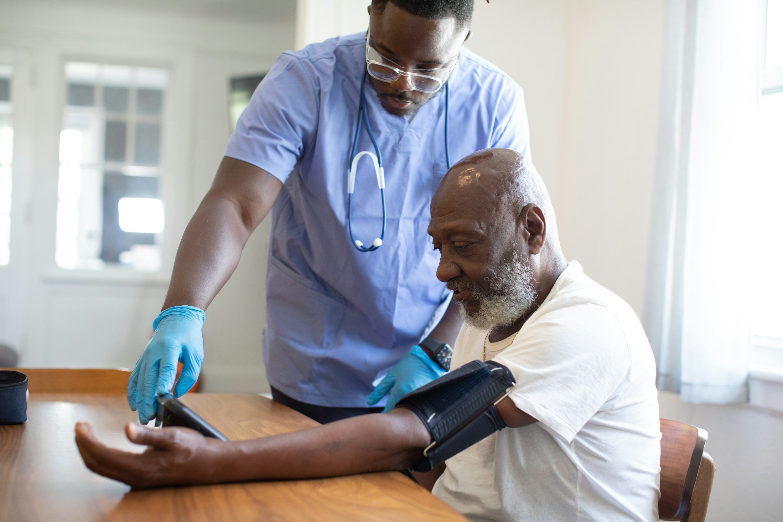 A healthcare professional, wearing glasses, a light blue scrub top, and a stethoscope around his neck, is attending to an elderly African American man with a long gray beard. The elderly man is seated at a wooden table, looking at a device on his wrist, possibly a blood pressure monitor, while the healthcare professional adjusts or checks the device.