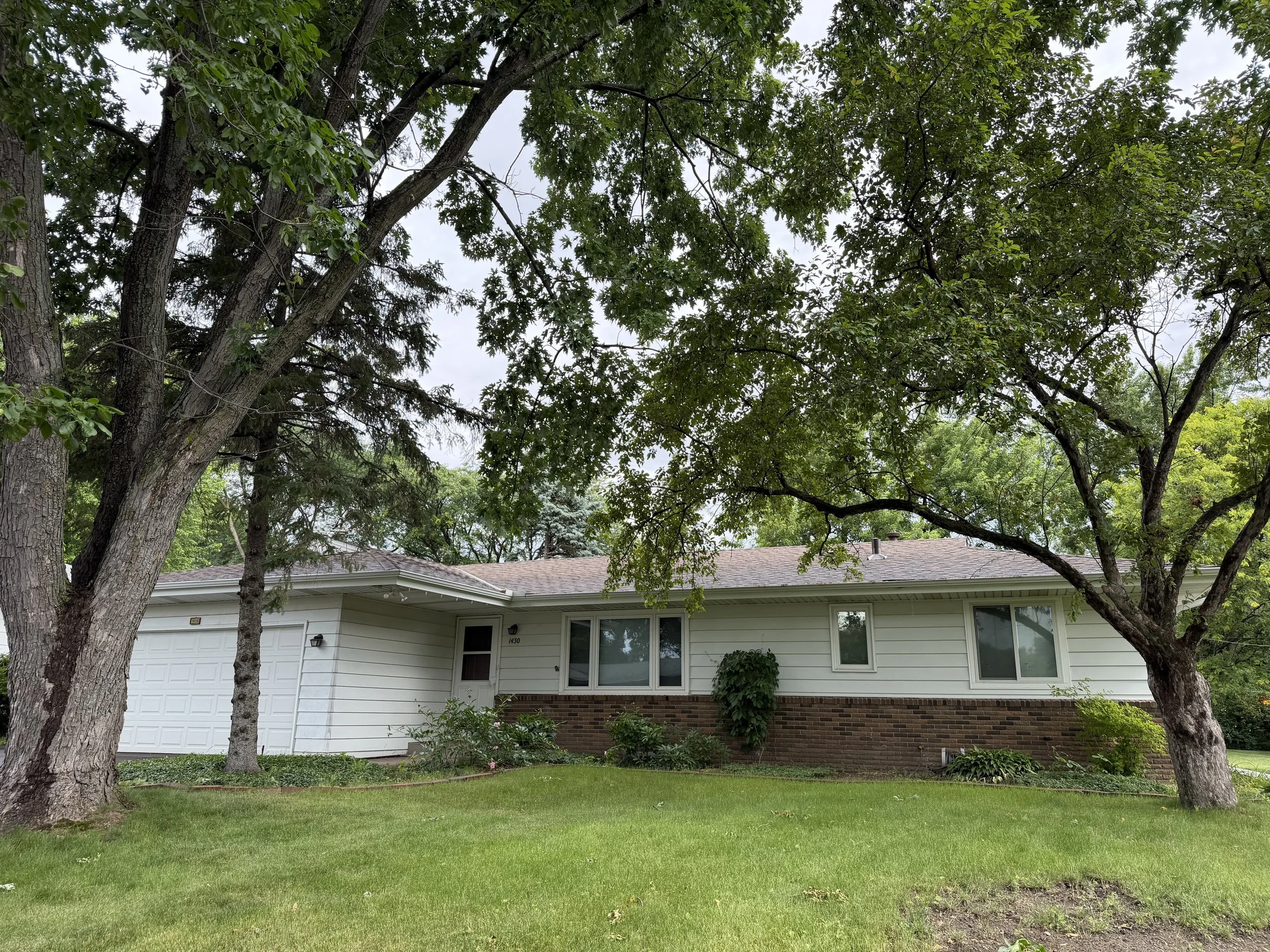 A suburban house with a white garage door, windows, and brick accents at the bottom, surrounded by green trees and grass.
