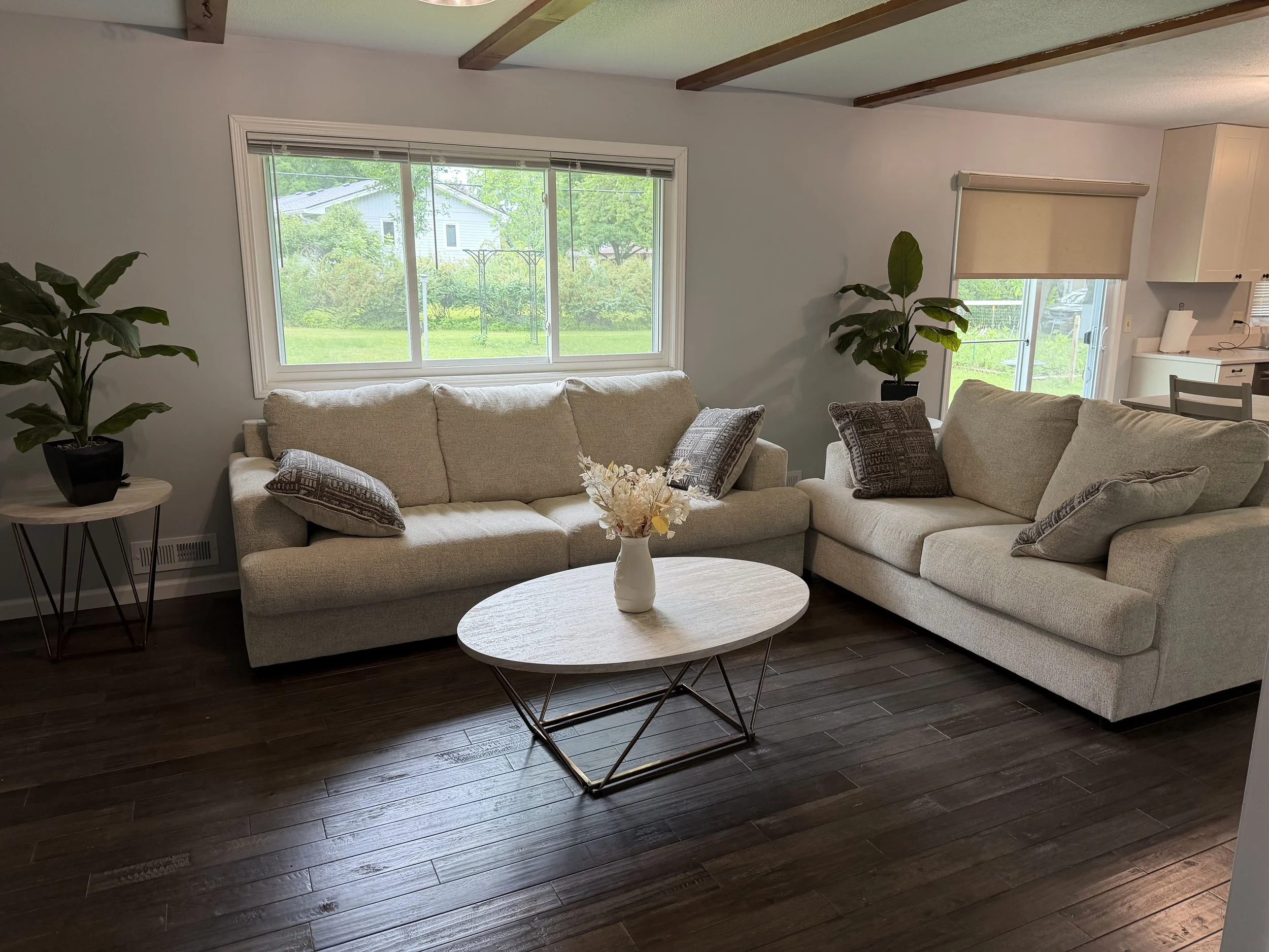 Living room with large window, beige couch with pillows, dark wood floor, coffee table with flower vase, side table with plant, kitchen area with window and light-colored cabinets.