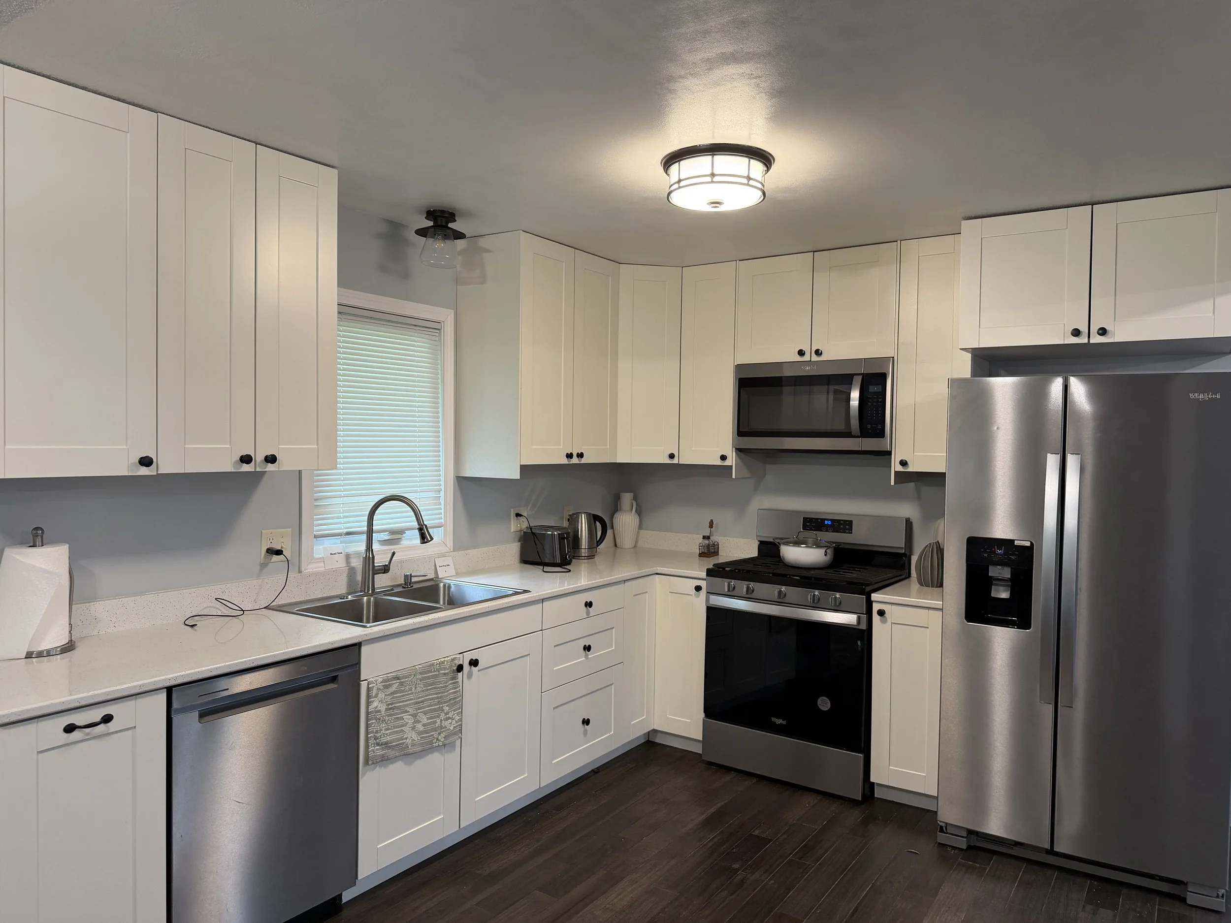 Modern kitchen with white cabinets, stainless steel refrigerator, microwave, and stove, dark wooden flooring, and a window with blinds.