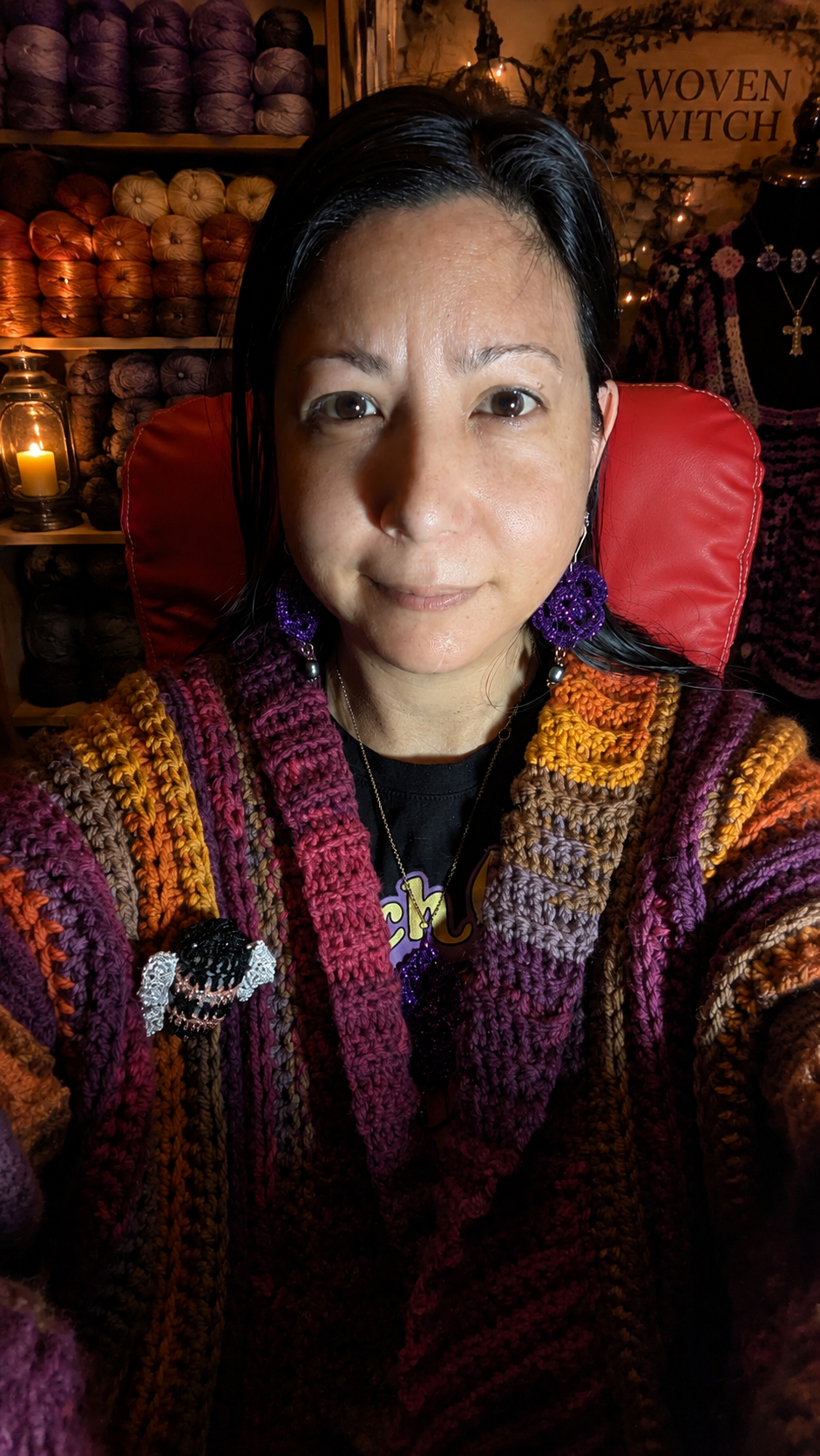 A woman with dark hair and wearing purple earrings takes a selfie in a craft store. She is wearing a multicolored, textured sweater with shades of purple, orange, yellow, and brown. The background shows yarn skeins on shelves and a sign saying 'WOVEN WITCH'.