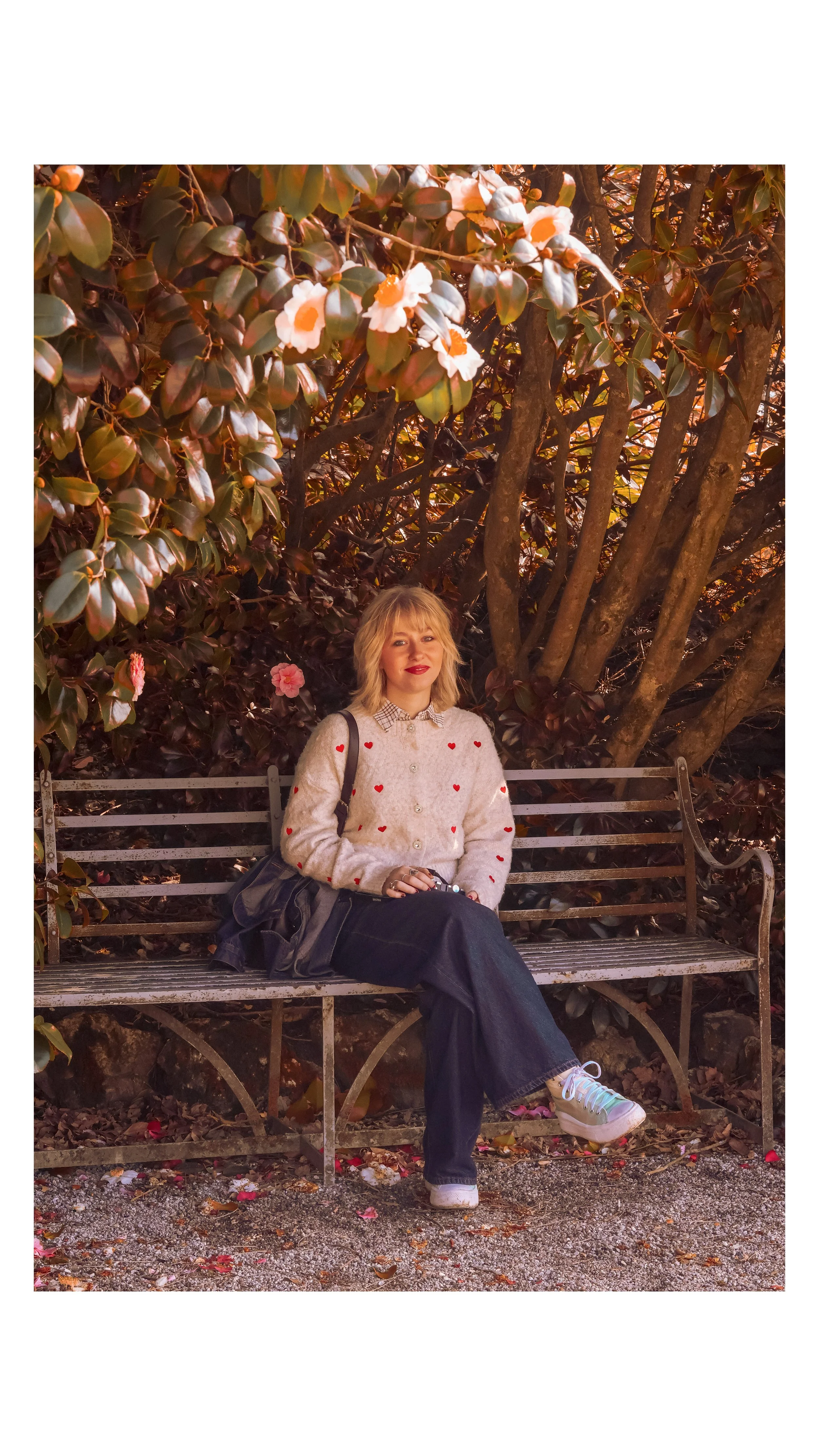 A woman sitting on a park bench surrounded by large shrubbery and trees with flowers and leaves. She is wearing a cream-colored cardigan with red hearts, dark pants, sneakers, and is holding a black bag.