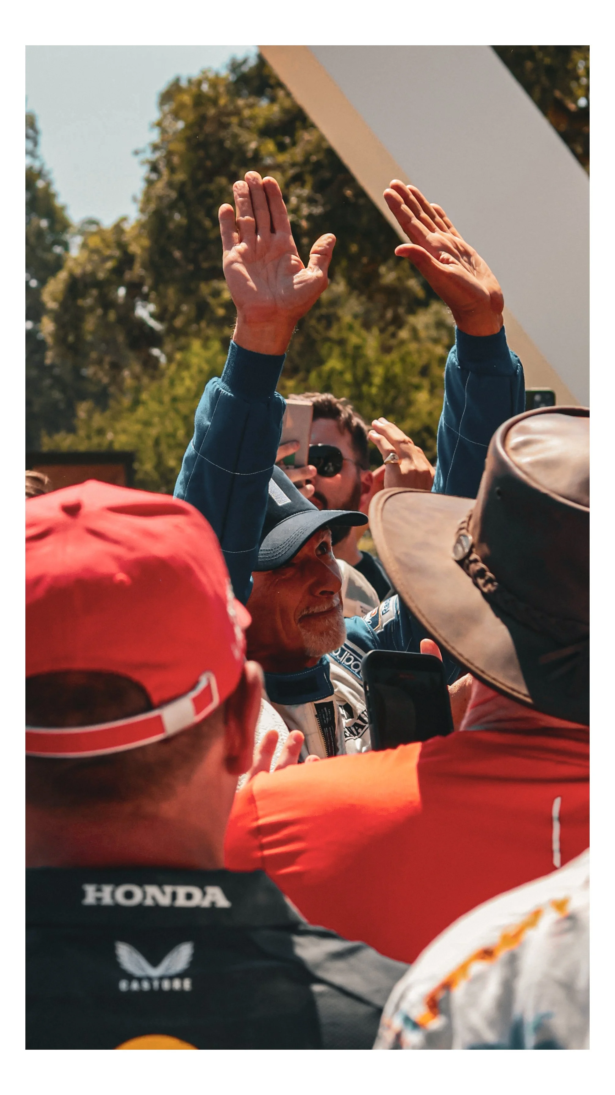A man with a cap and racing suit, surrounded by fans, raising his hands and smiling during an outdoor event with trees in the background.