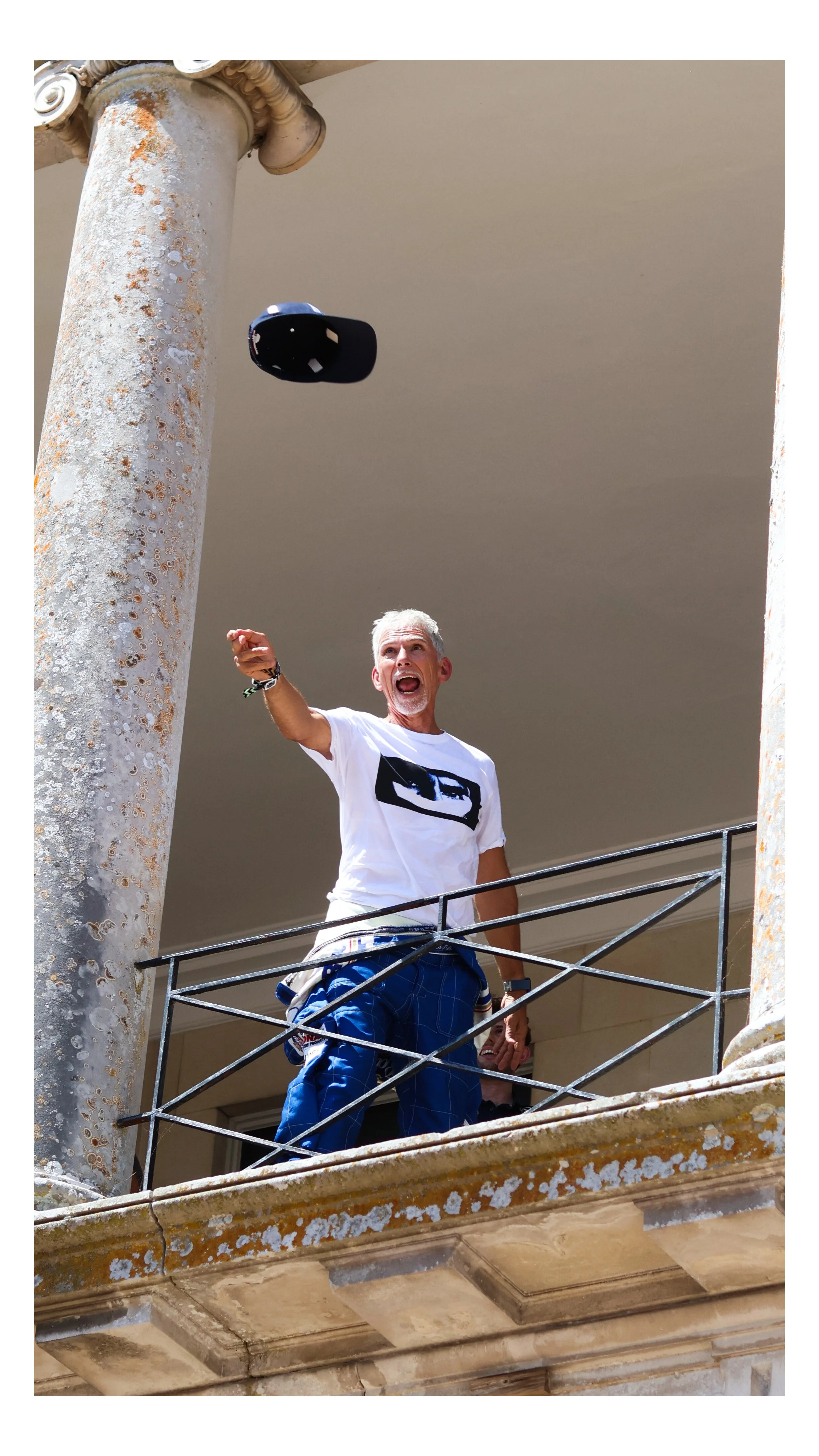 A man with gray hair and a beard is standing on a balcony railing, shouting or calling out, wearing a white t-shirt and blue pants. There is a cap flying in the air overhead, and the man appears to be pointing or gesturing with his right hand. The balcony has a weathered stone pillar and rusted metal railing.