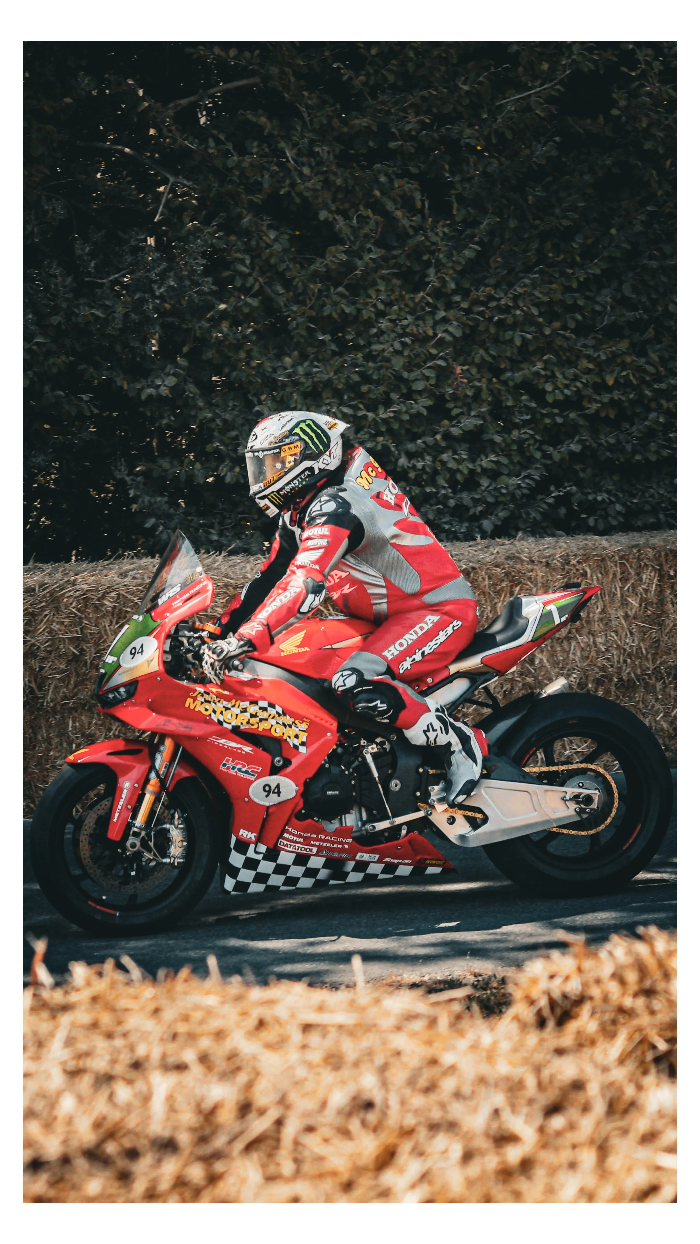 Motorcycle racer in red racing suit riding a red Honda motorcycle on a paved road with dry brush and foliage in the background.