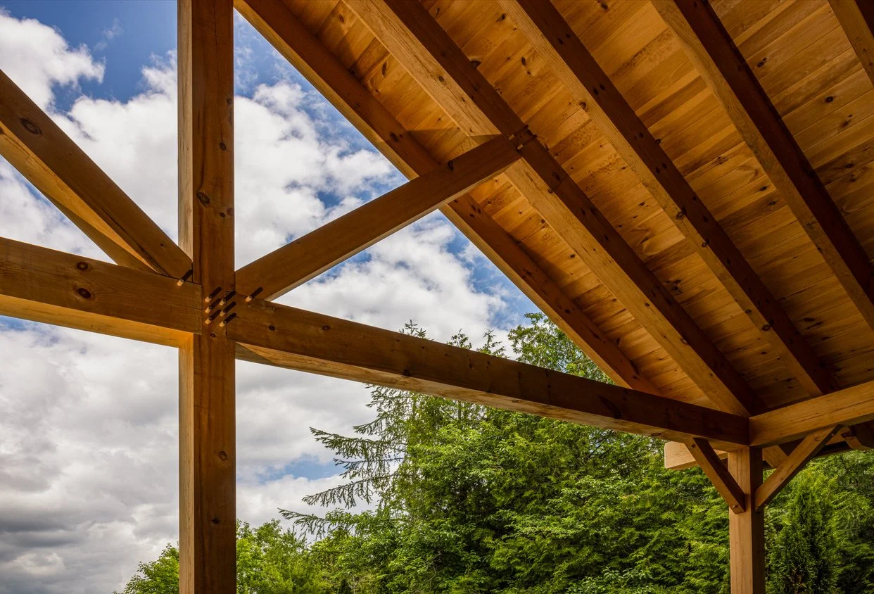 View of a construction with wooden beams and roof, looking up at the sky and clouds, with green trees in the background.