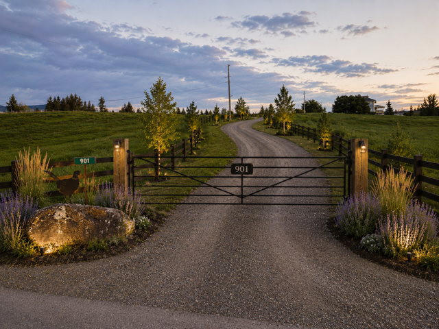 A gravel driveway with a black metal gate leading into a green field, surrounded by lit outdoor lights, small trees, and purple and yellow flowers at dusk.