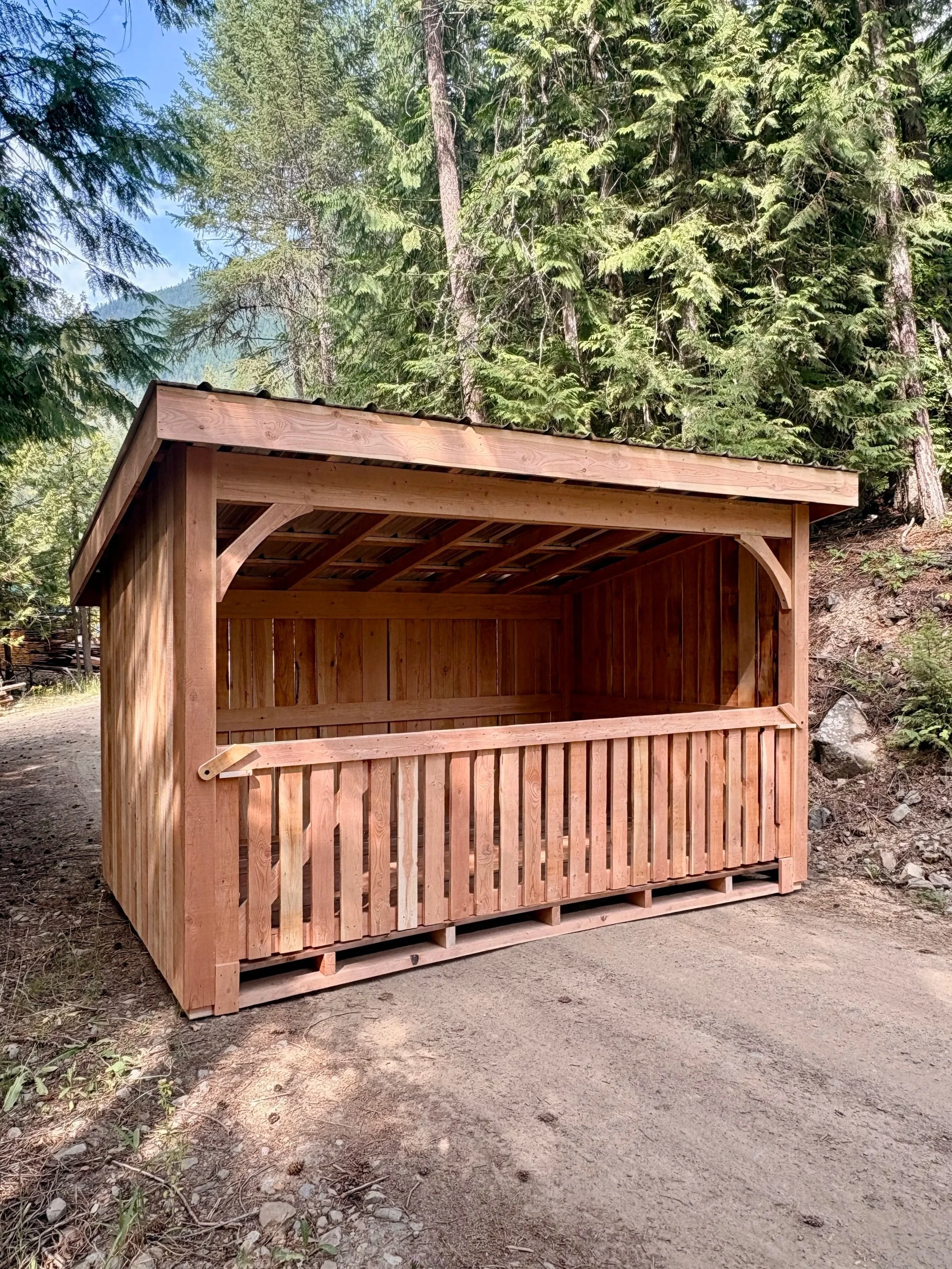 A wooden shelter with a slanted roof situated on a dirt path in a forested area with tall green trees.