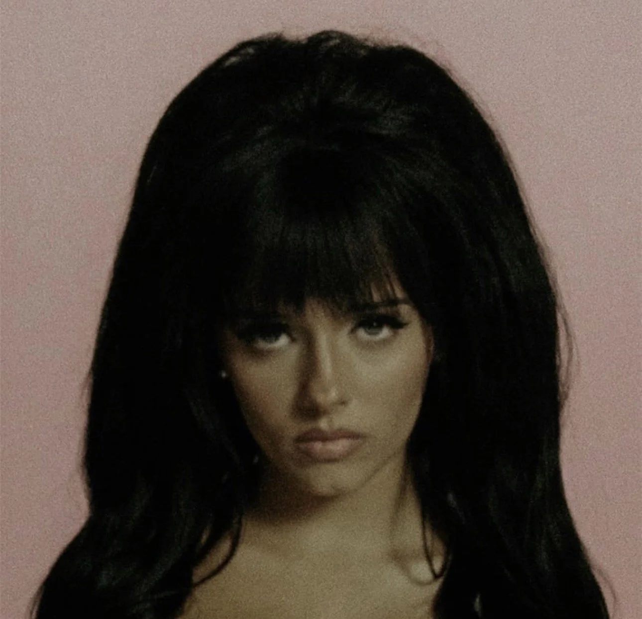Close-up of a woman with dark, voluminous hair and makeup against a pink background.