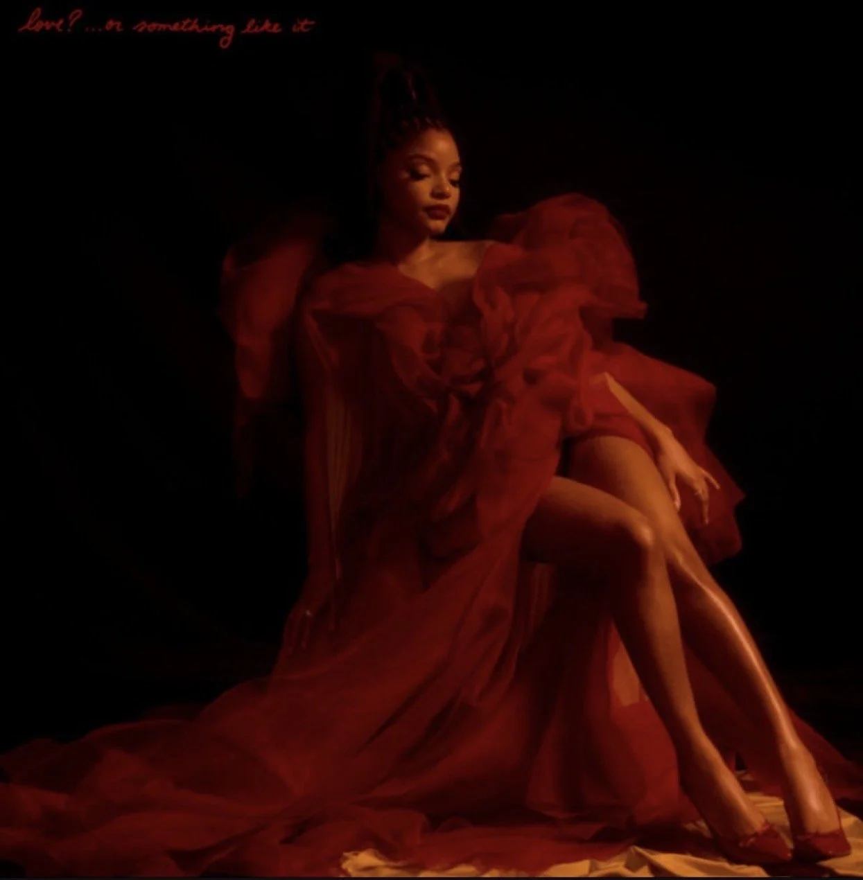 A woman sitting on the floor dressed in a flowing, deep red gown with layered, ruffled sleeves, against a dark background, with soft lighting highlighting her face and legs.