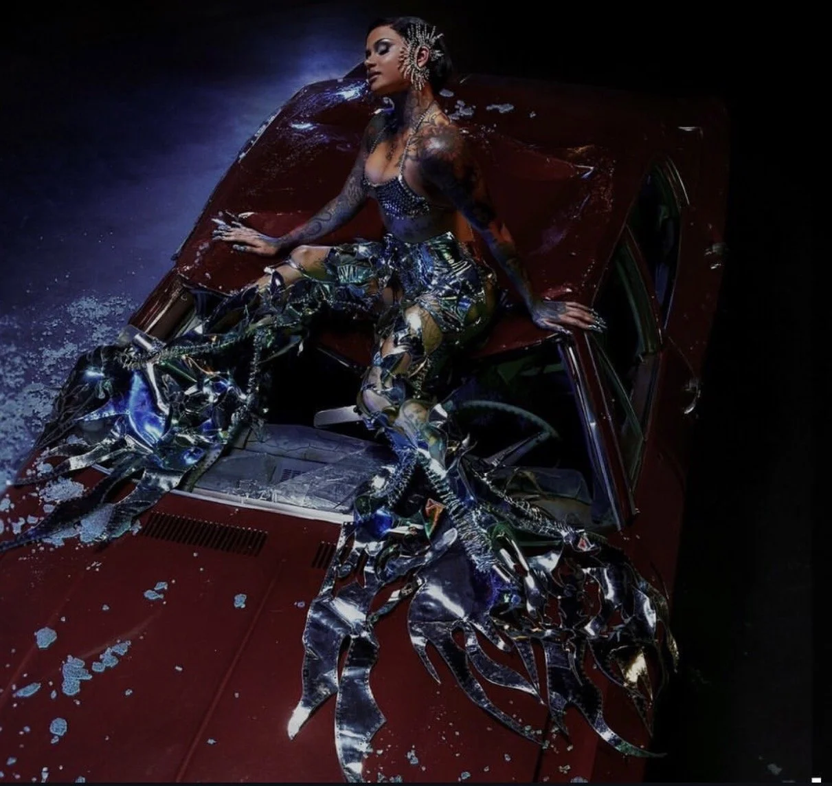 A woman with tattoos sitting on the hood of a red vintage car with broken glass and water around her, wearing futuristic metallic and colorful clothing and accessories, in a dark setting.