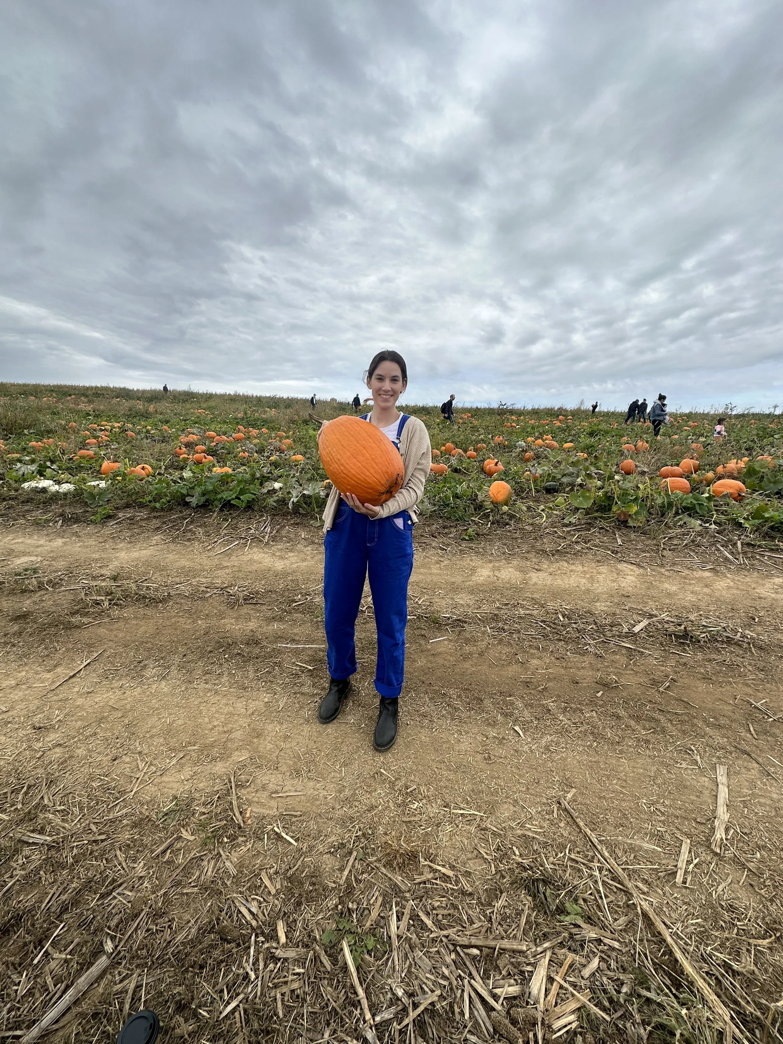 A smiling woman holding a large orange pumpkin in a pumpkin patch under a cloudy sky, with several people in the background.