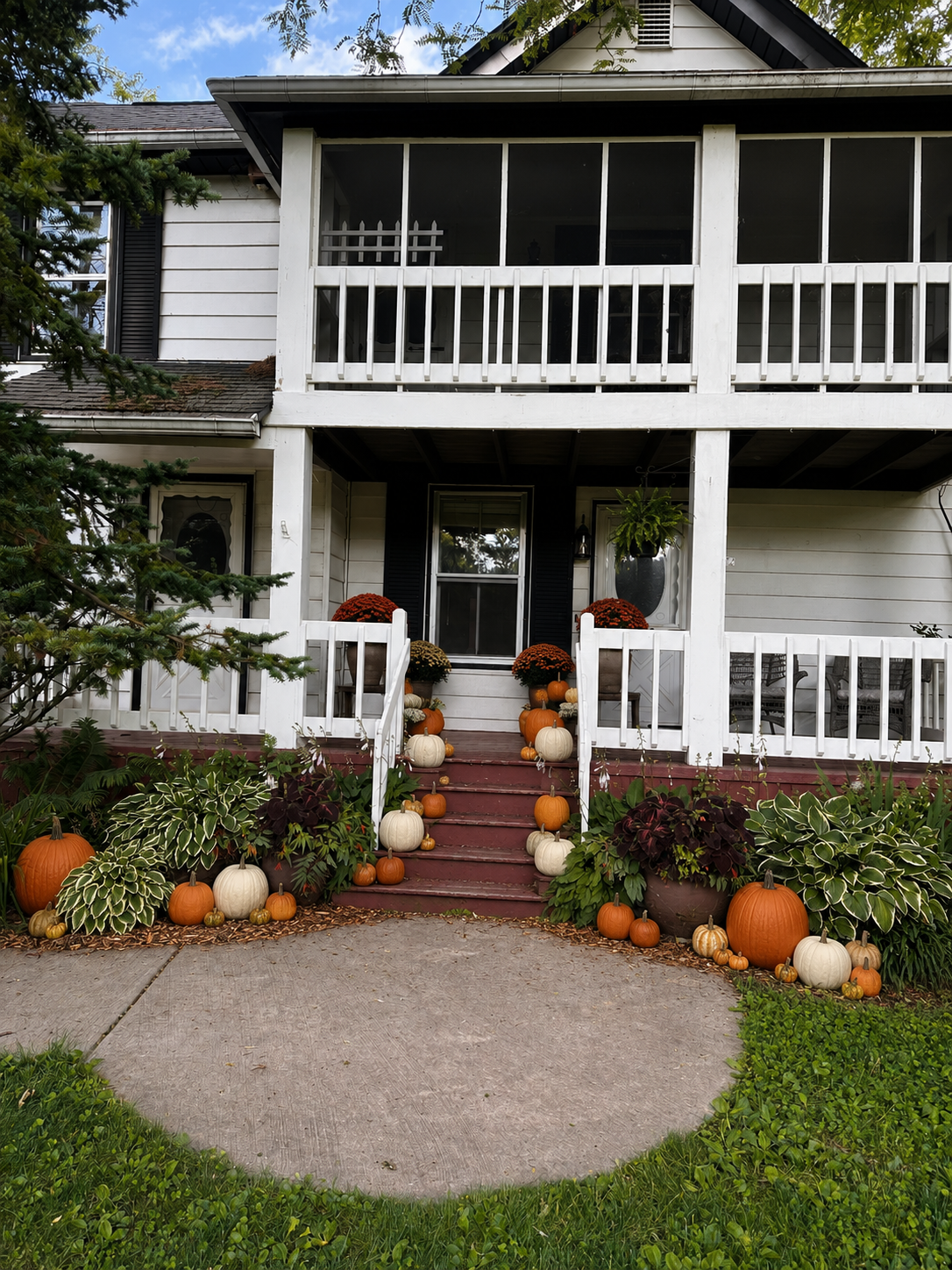 Front porch of a house decorated for fall with pumpkins and mums, with a walkway and greenery in the foreground.