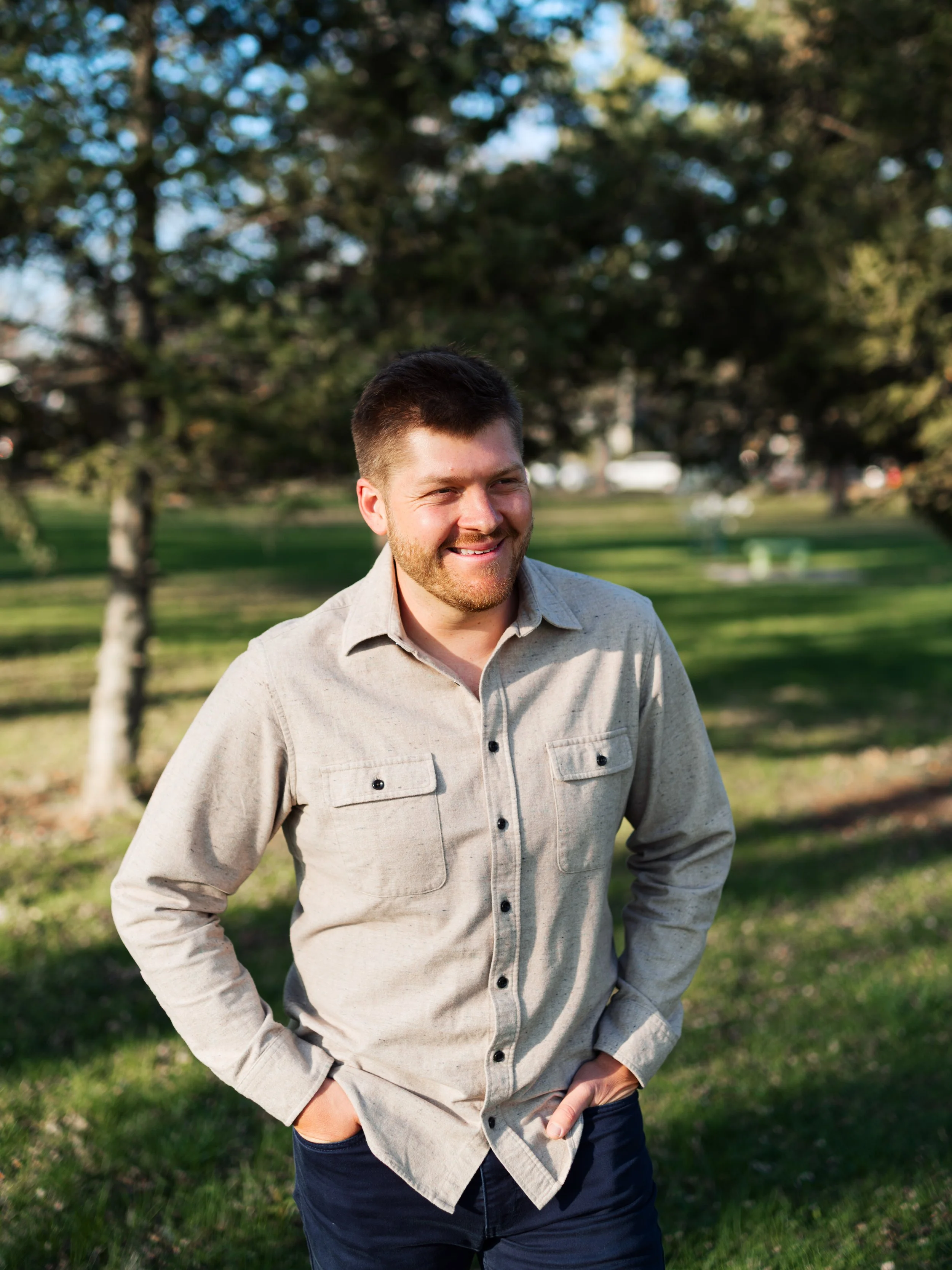 Smiling young man with short dark hair and beard, wearing a beige button-up shirt and dark jeans, standing outdoors in a park with green grass, trees, and a sunny sky.