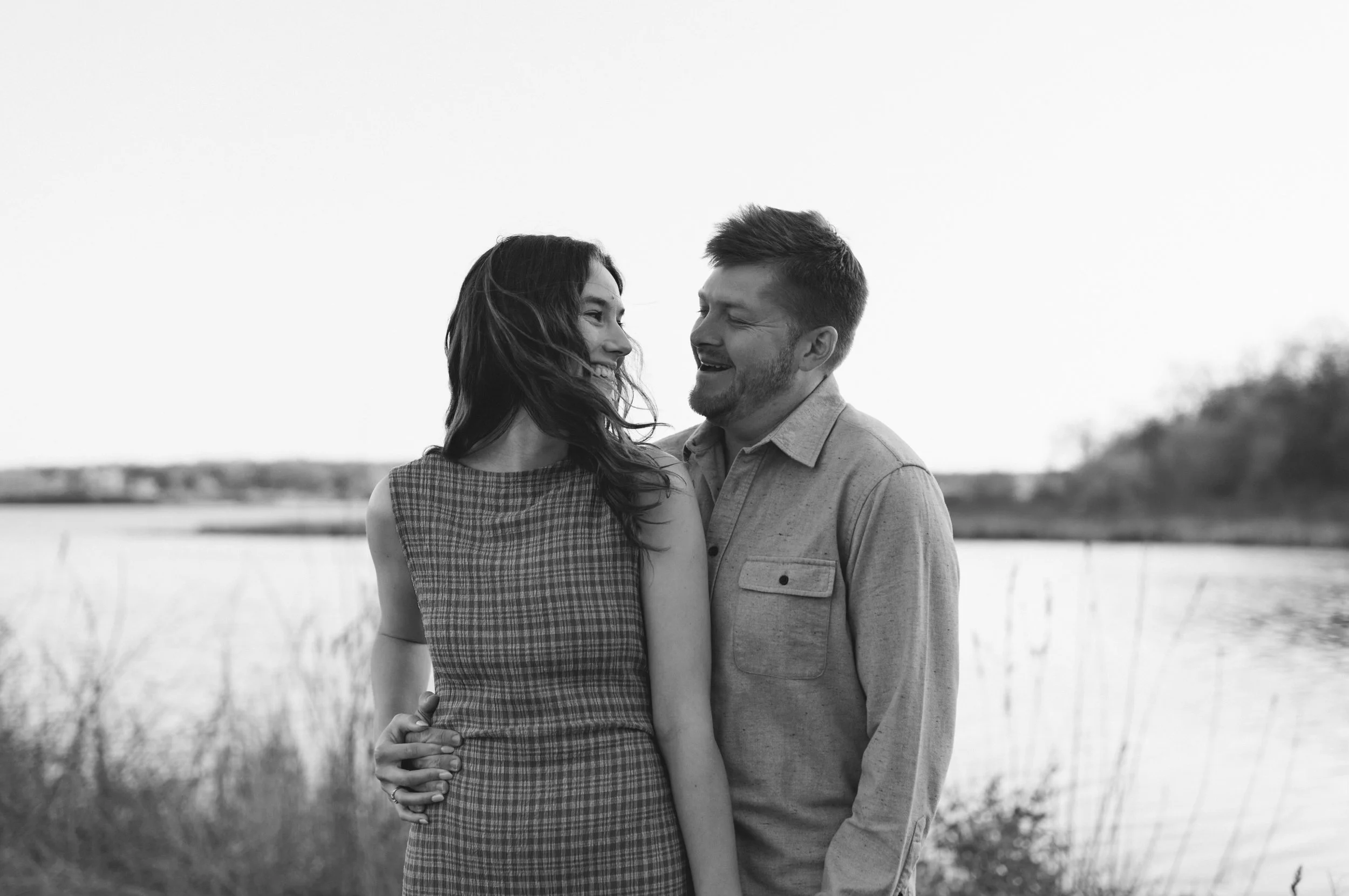 A black and white photo of a couple standing outdoors near a body of water with trees and grass in the background. The woman has long wavy hair and is wearing a sleeveless dress, while the man has short hair, a beard, and is wearing a button-up shirt. They are smiling and looking at each other affectionately.