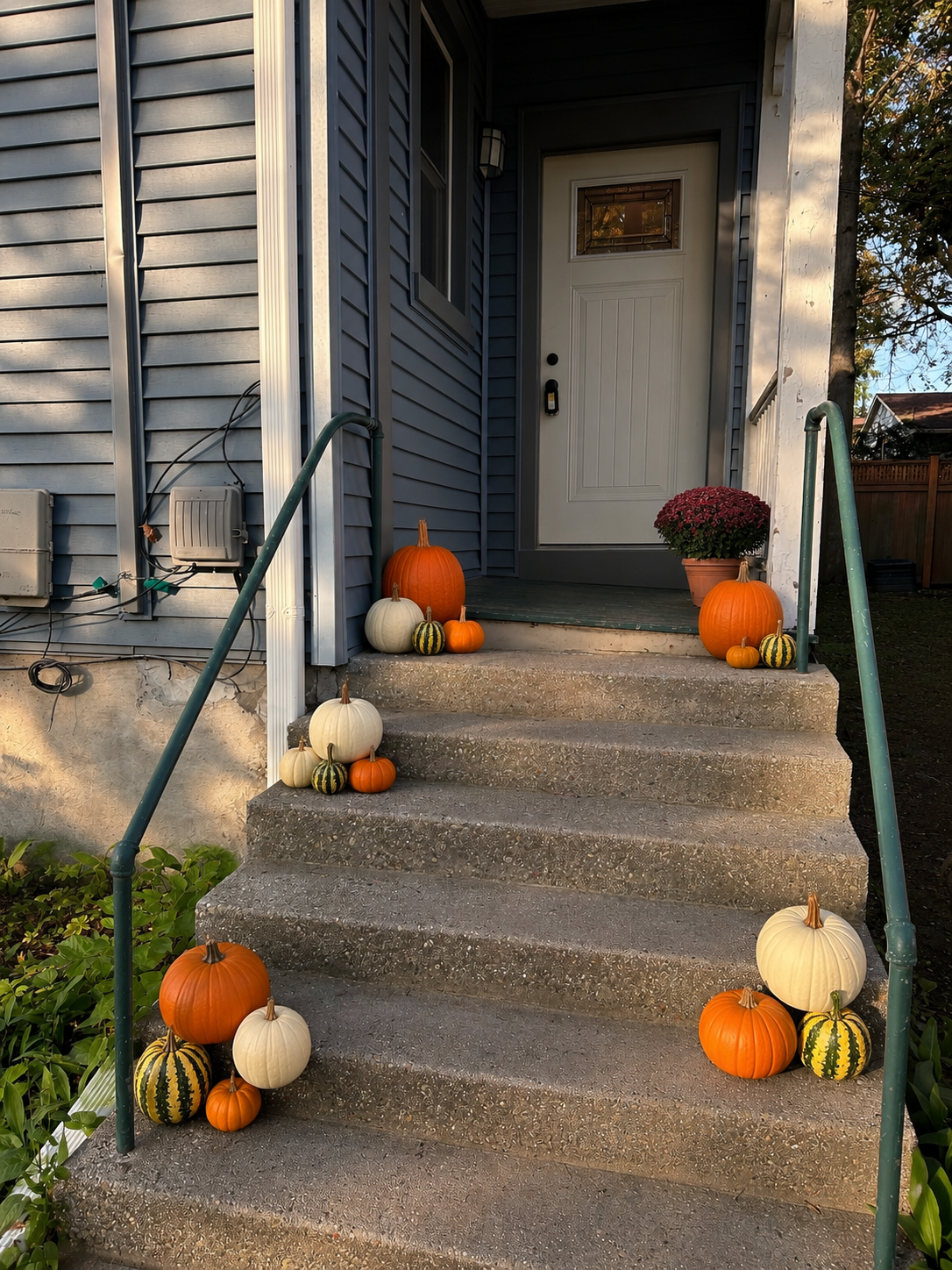 Front porch stairs decorated with pumpkins of various sizes and colors, including orange, white, green-striped, and yellow-striped pumpkins. A potted plant with pink flowers is on the right side of the porch. The house has blue siding and a white door with a window.