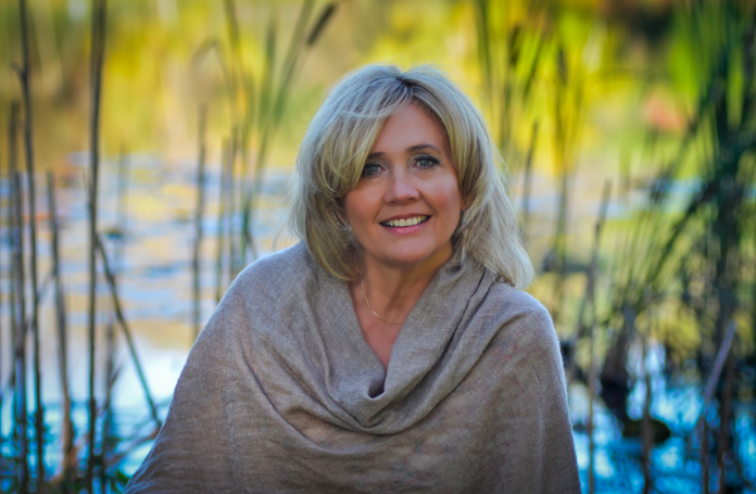 A woman with blonde hair smiling outdoors in front of a pond with reeds and green foliage.