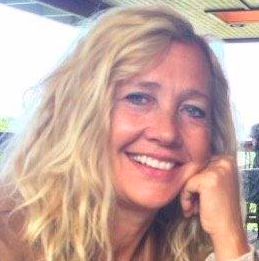 A woman with long, wavy blonde hair smiling and resting her chin on her hand in an outdoor setting