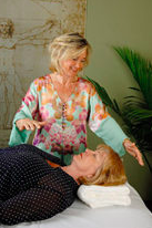 A woman standing next to a bed, gently touching a sleeping woman's forehead, in a room with a green plant and light-colored wall.