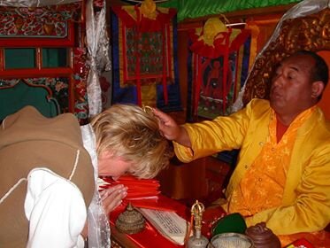 A person with blond hair bowing or praying in front of a man in traditional yellow and orange robes inside a decorated, colorful setting.