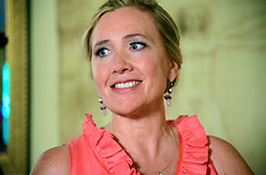 A smiling woman with blonde hair wearing a coral-colored sleeveless top, standing indoors with a light-colored wall in the background.