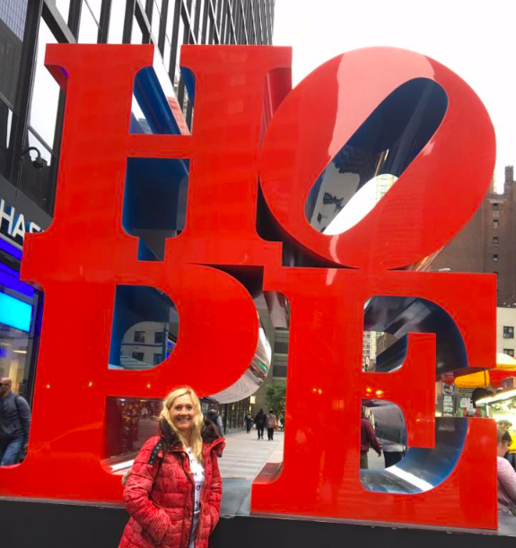 Person standing in front of large red "LOVE" sculpture in an urban area, with people and buildings in the background.