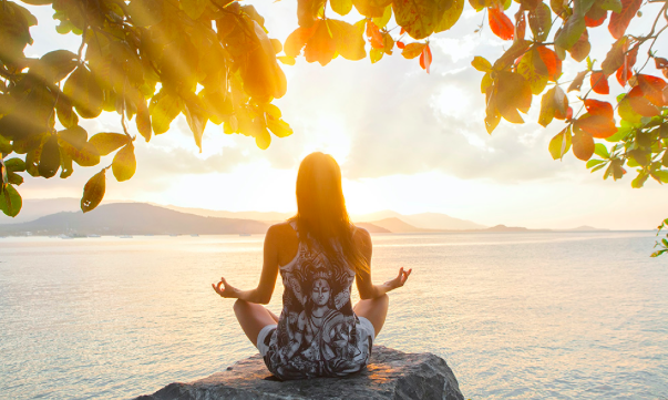 A woman sitting cross-legged on a rock near the water, meditating during sunset, with trees and mountains in the background.