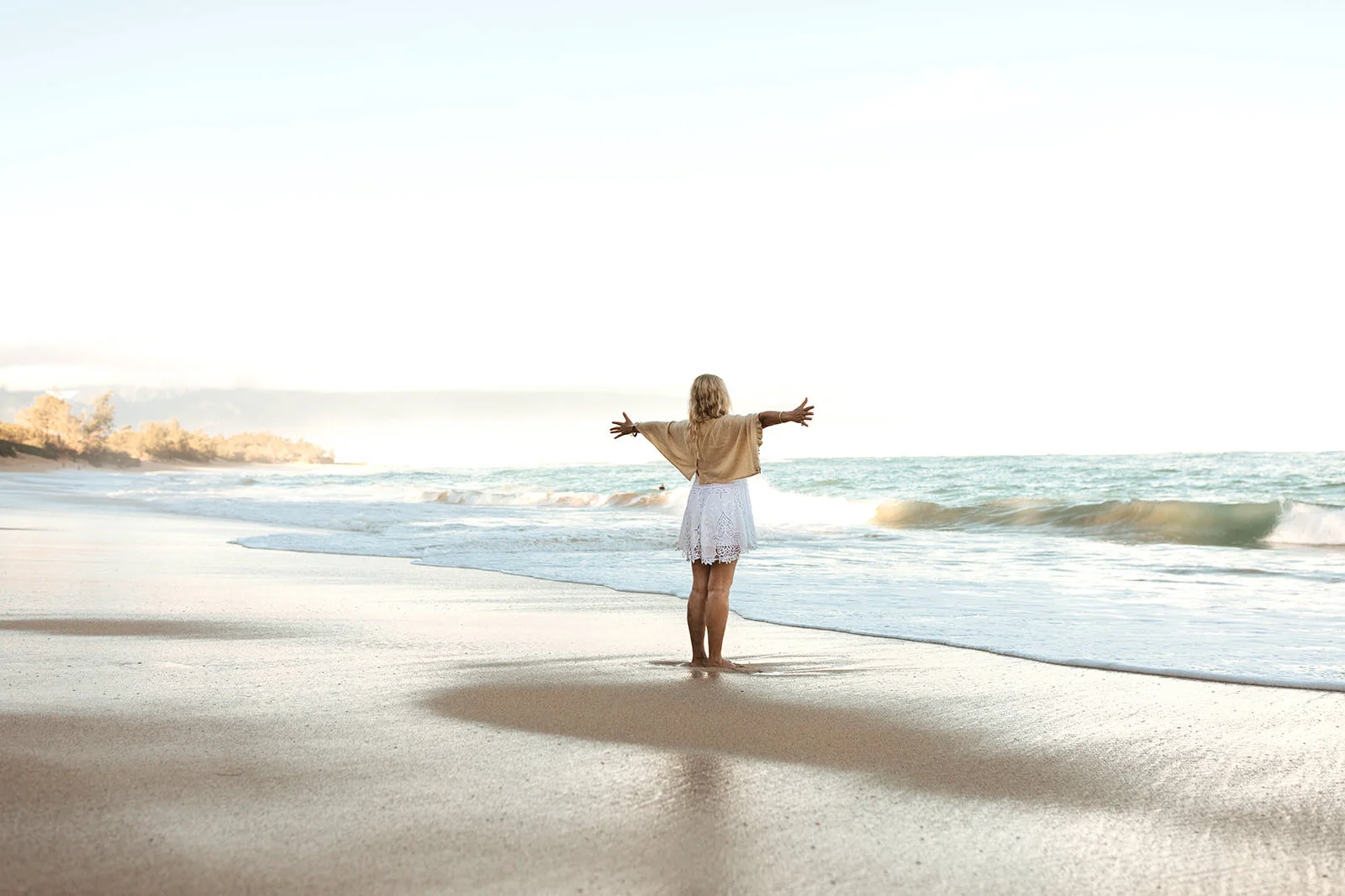 A woman stands on a sandy beach with her arms outstretched, facing the ocean, with waves gently crashing and a distant shoreline with trees in the background.