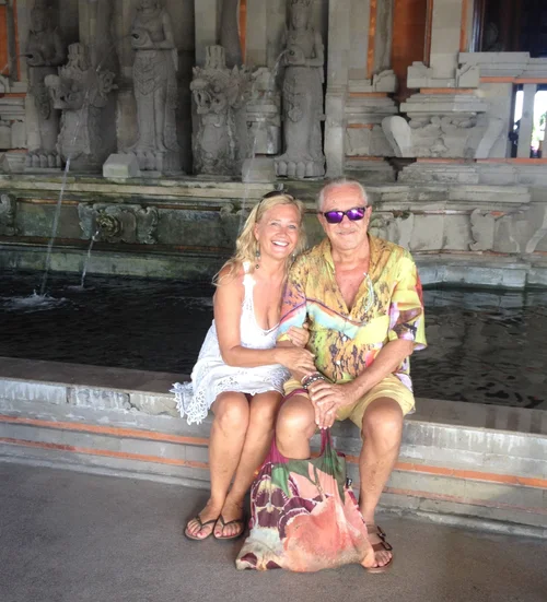 A smiling woman and man sitting together on a bench in front of a water fountain with decorative stone sculptures, dressed casually in summer clothing.