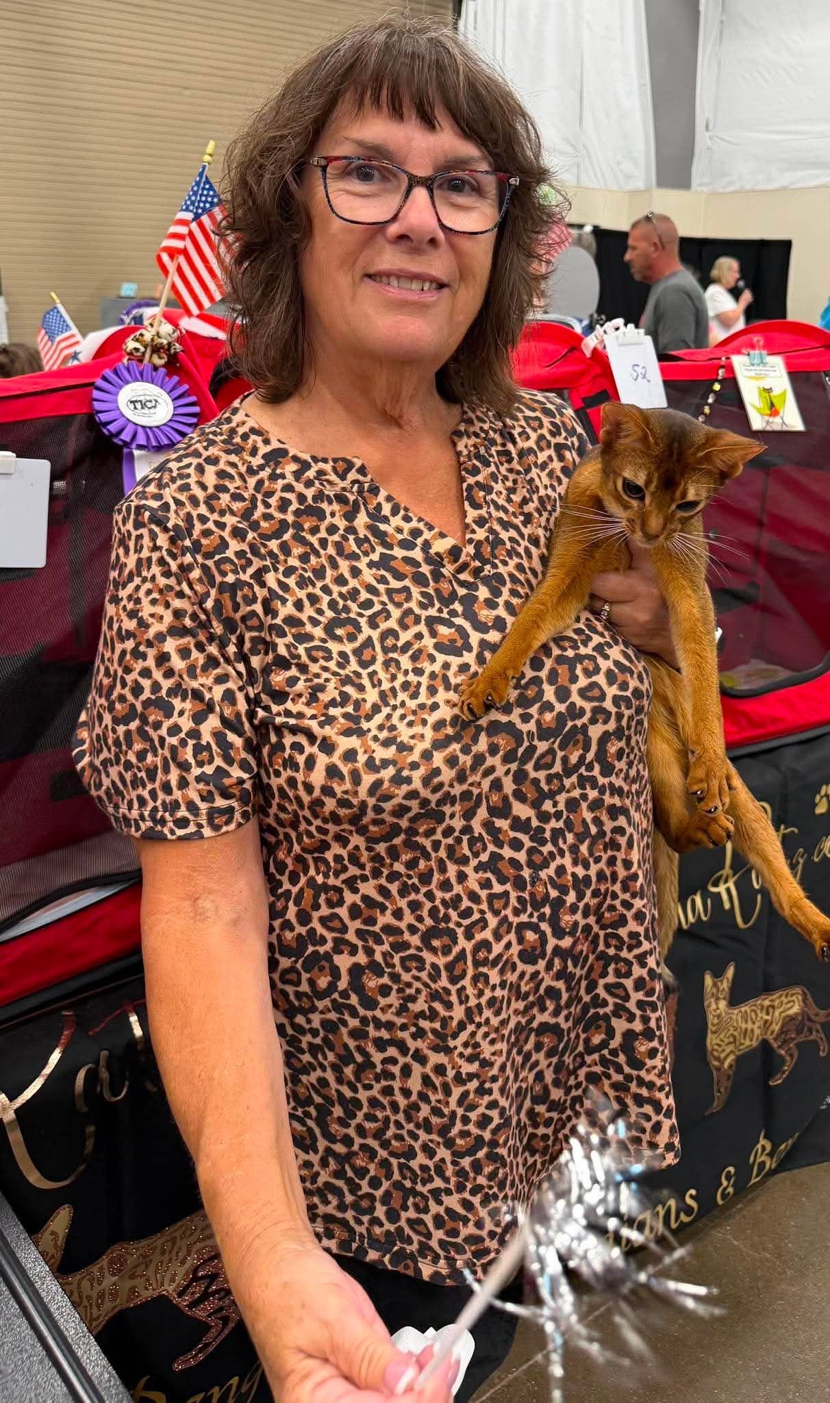 A woman with glasses and a leopard print shirt holding a brown cat at an indoor event with American flags and awards in the background.