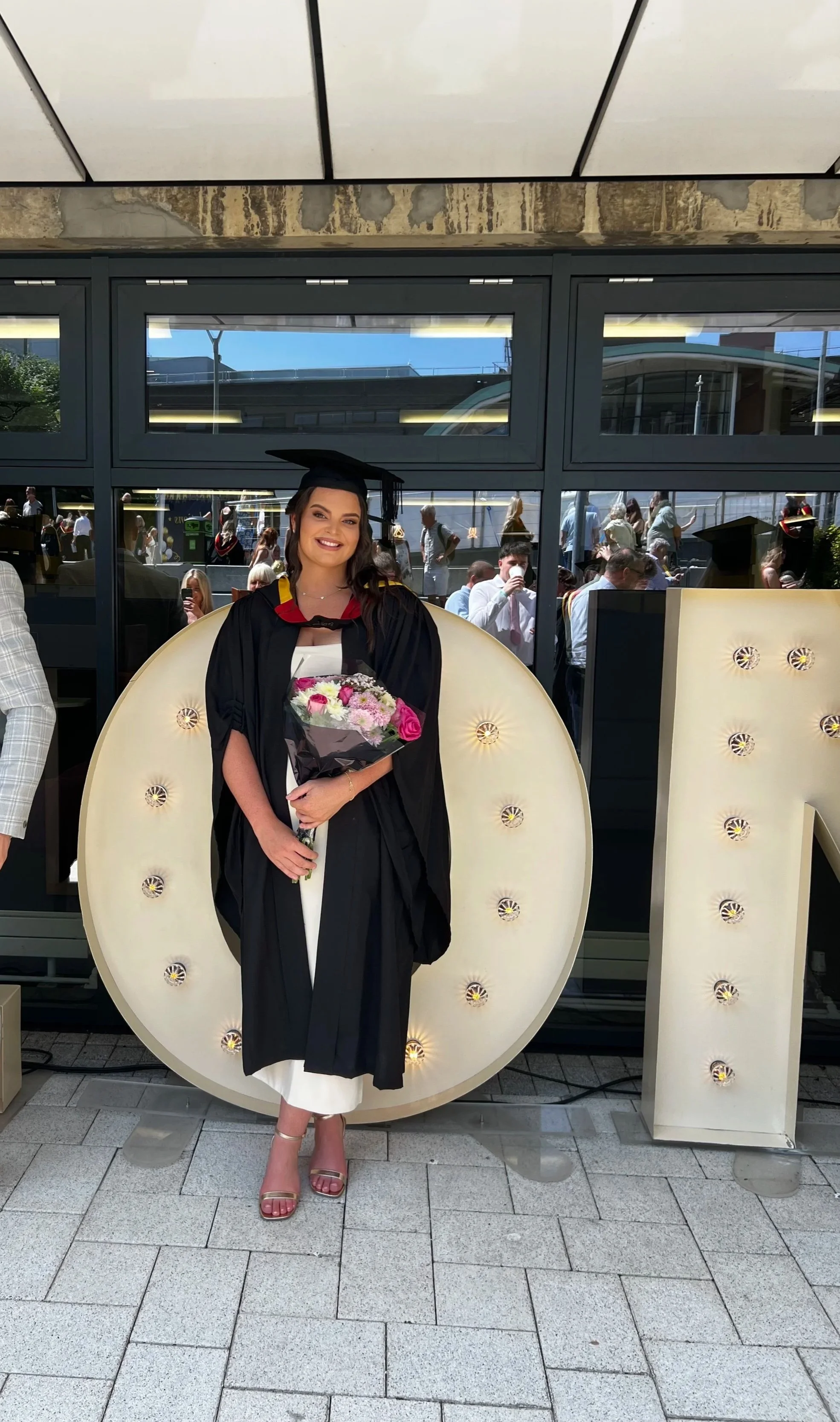 A woman in a graduation cap and gown holding a bouquet of flowers, standing in front of large illuminated letters outdoors with a crowd of people reflected behind glass windows.