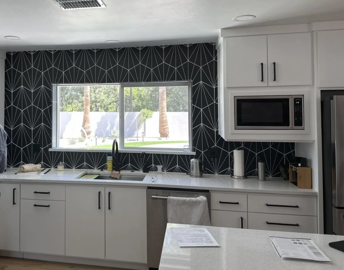 Modern kitchen with white cabinets, black geometric backsplash, stainless steel appliances, window showing trees outside, and kitchen counter with paper towels, kettle, and papers.