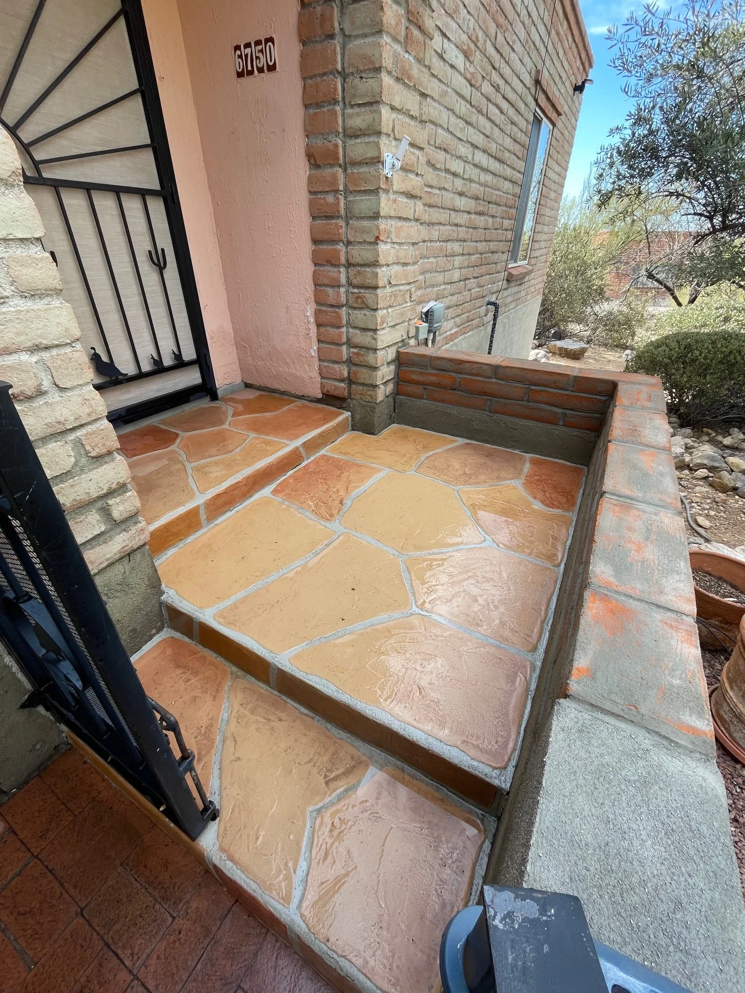 Front porch area with multicolored stone tiles, brick and stucco walls, security door with decorative peacock and spear design, house number 6750, security camera, electrical meter, and outdoor greenery with trees and bushes.