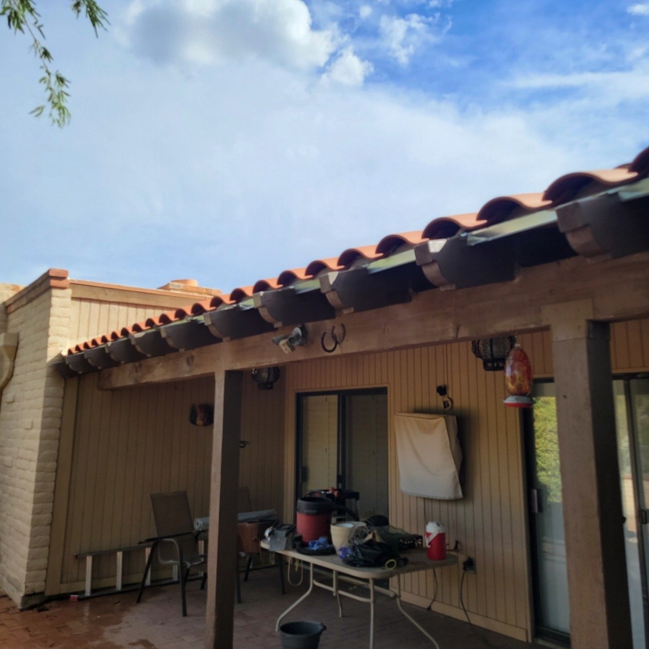Back porch of a house with a wooden roof structure, hanging lanterns, and a table with various items including a red cooler and a black vacuum.