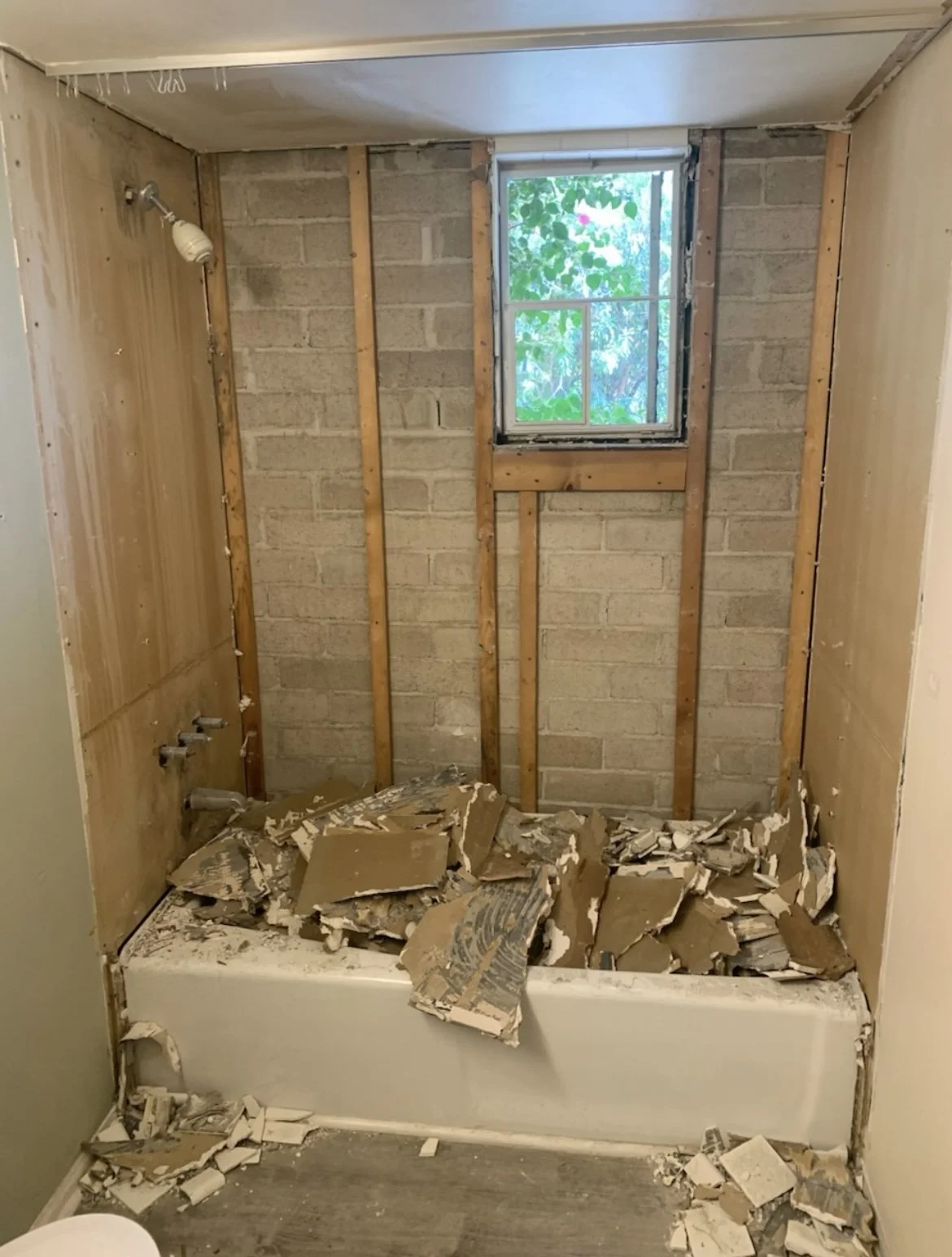 Bathroom in the process of renovation with removed tiles, exposed wall studs, a small window, and debris on a bathtub.