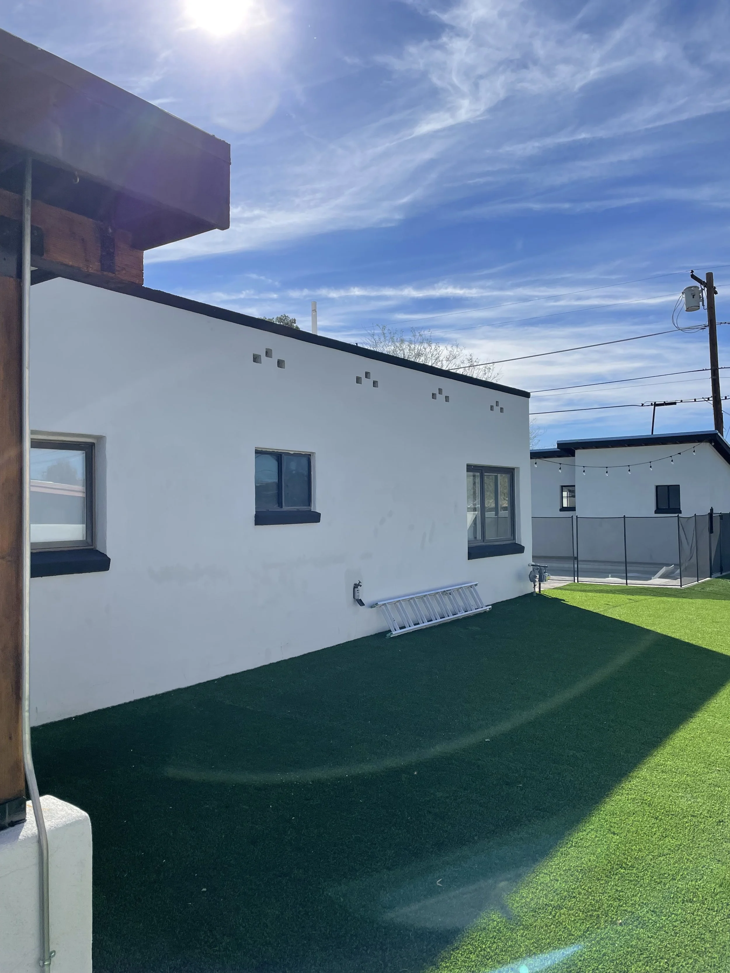 A backyard with artificial green grass, modern white houses with black window frames, and a clear blue sky with wispy clouds. There's a ladder on the ground and string lights hanging between the houses.