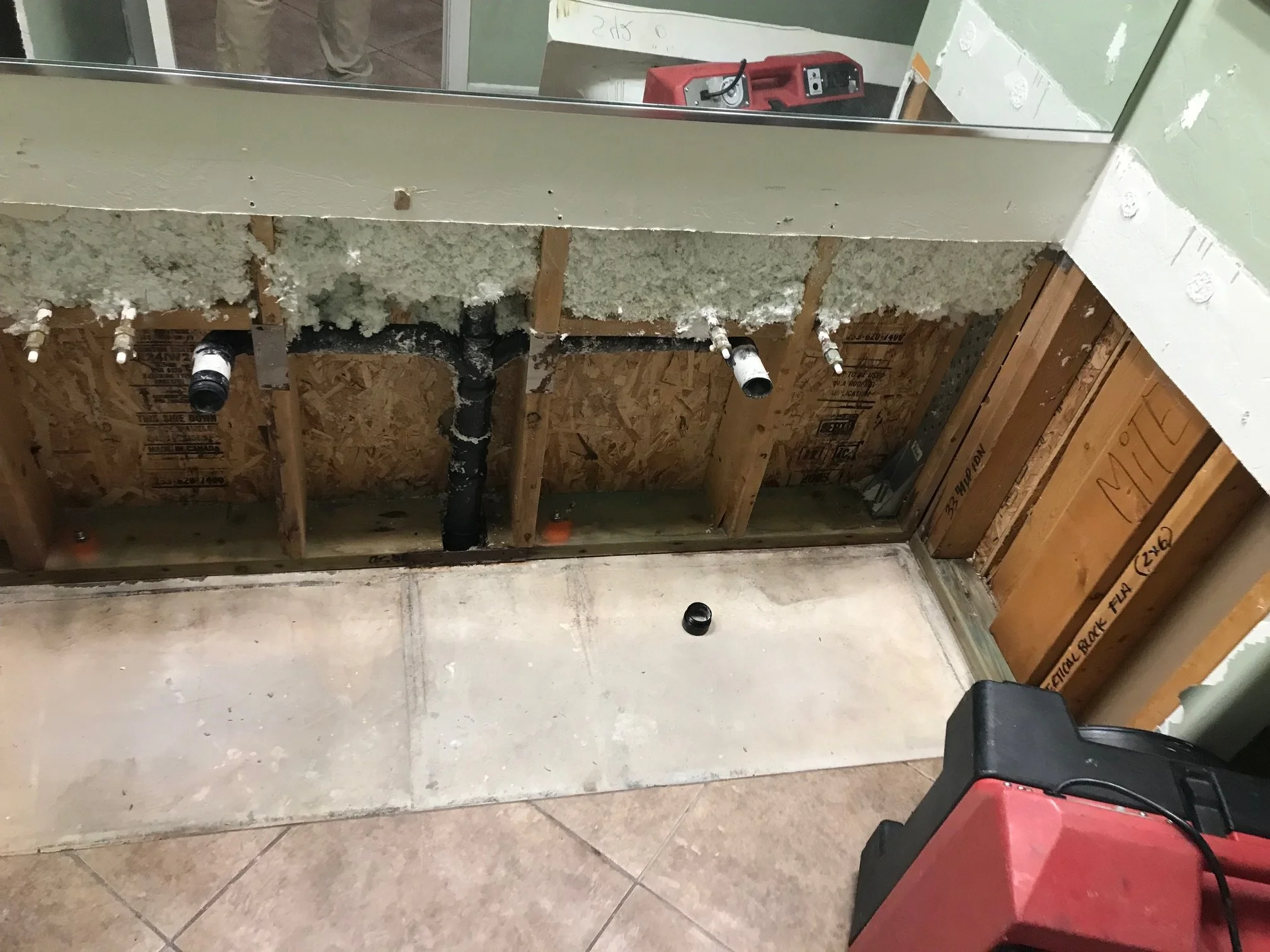 Construction site showing a partially built wall with insulation material. There are pipes and black fittings visible, and construction tools like a red power tool are present. The area has exposed studs and drywall in progress.