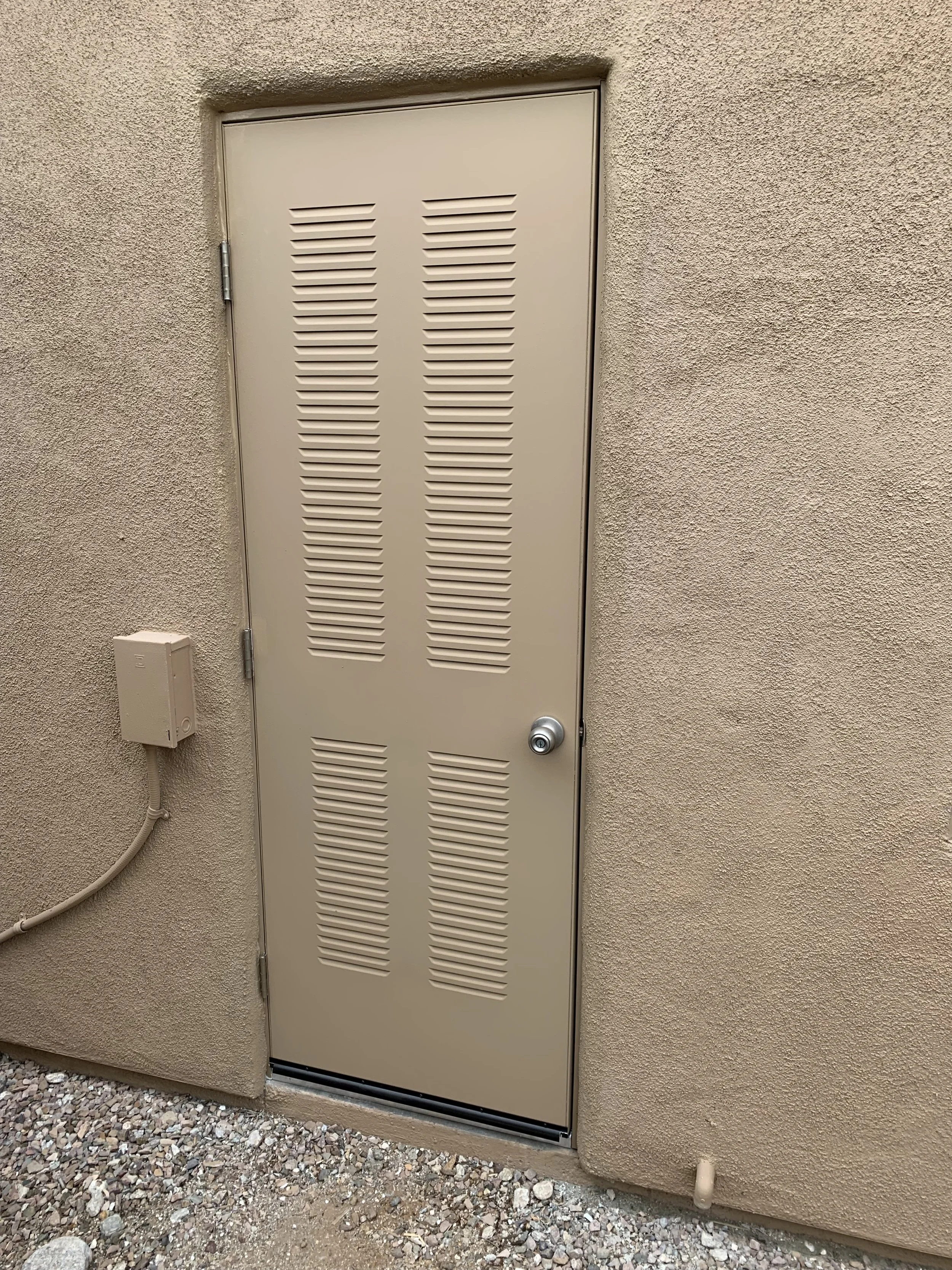 A beige outdoor door with ventilation slats, set in a textured beige wall, with a round doorknob, a small electrical box on the left, and a gravel ground below.