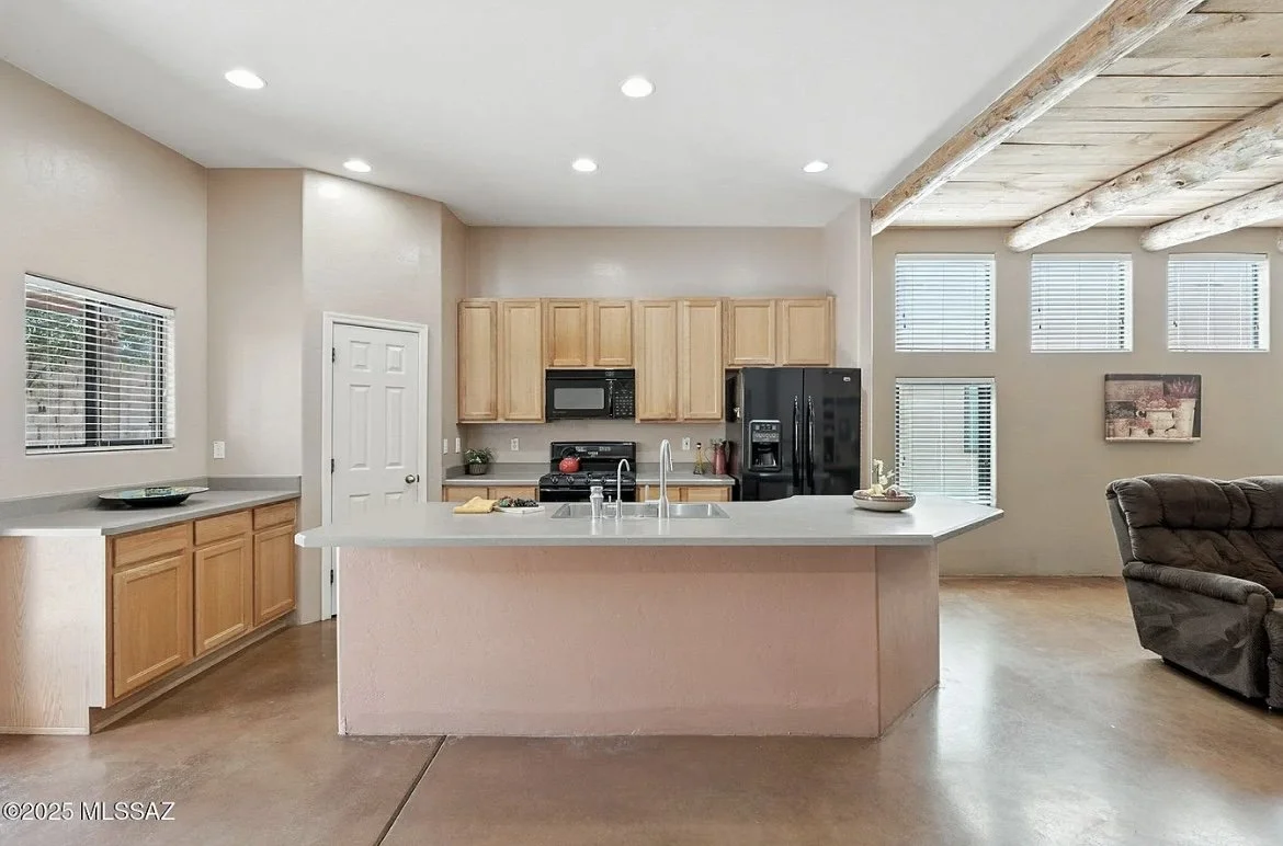 Kitchen with light wood cabinets, black appliances, and a large central island with a double sink, open-style living area with multiple windows and a reclining chair, ceiling beams, and recessed lighting.