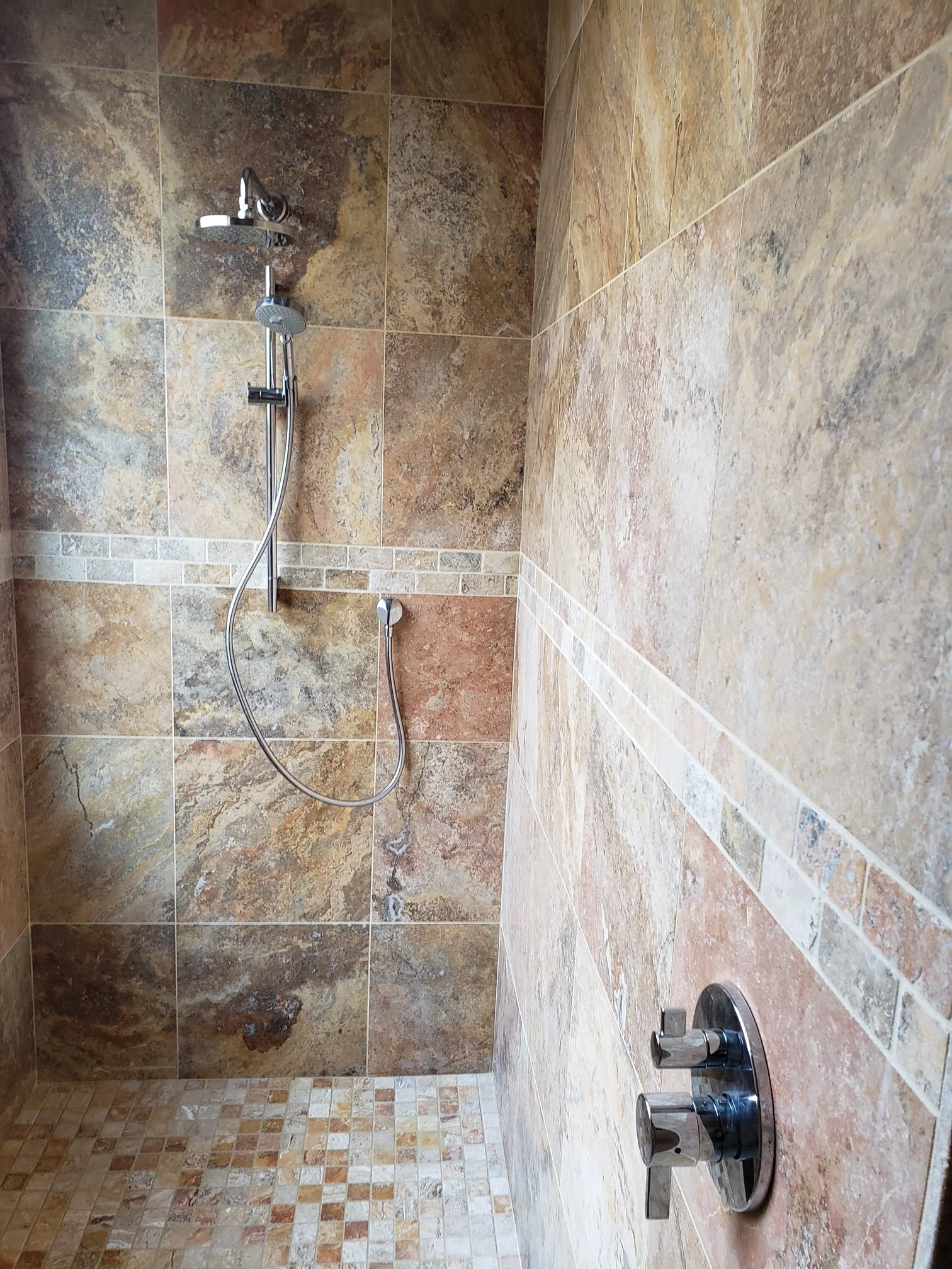 A bathroom shower with multi-colored stone tiles, a handheld showerhead, and a drain on the floor.