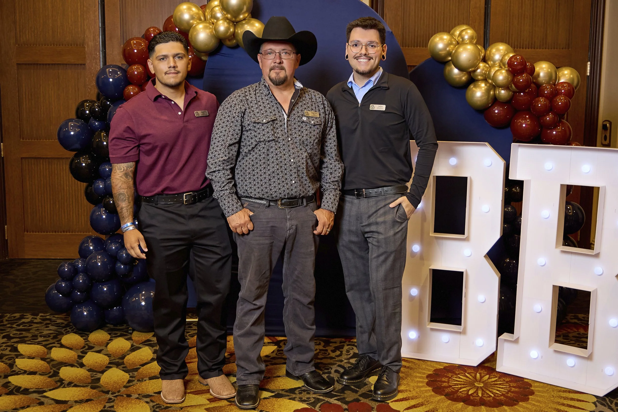 Three men standing in front of a decorative backdrop of balloons and a letter 'K' with lights. They are at an indoor event or celebration.