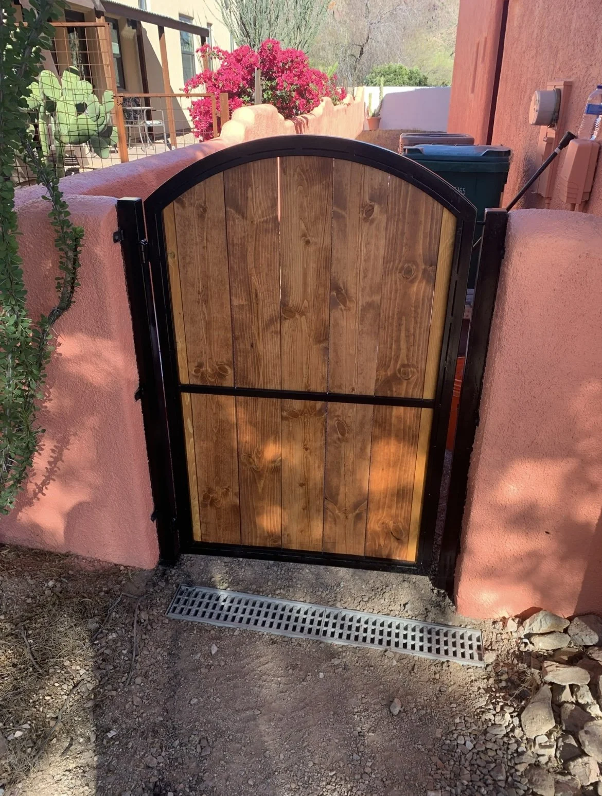 A wooden gate with a black metal frame, installed between two pink stucco walls. In the background, there are pink flowering bushes, a potted plant, and trees. A drainage grate is visible on the ground in front of the gate.