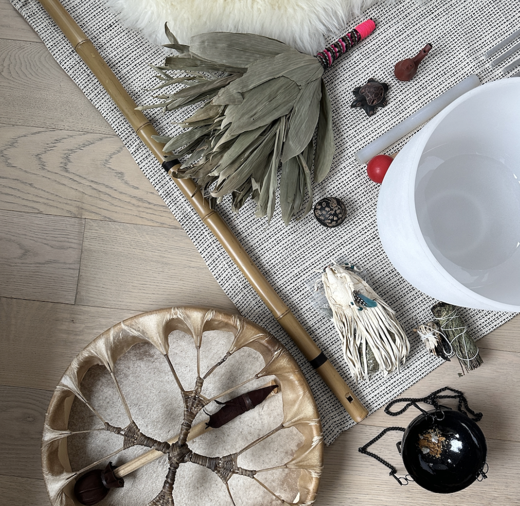 Collection of Native American or indigenous sound instruments, including a chakapa, a drum, a medicine bag, sound bowl, and a black incense burner, arranged on a woven fabric surface with a wooden floor underneath.