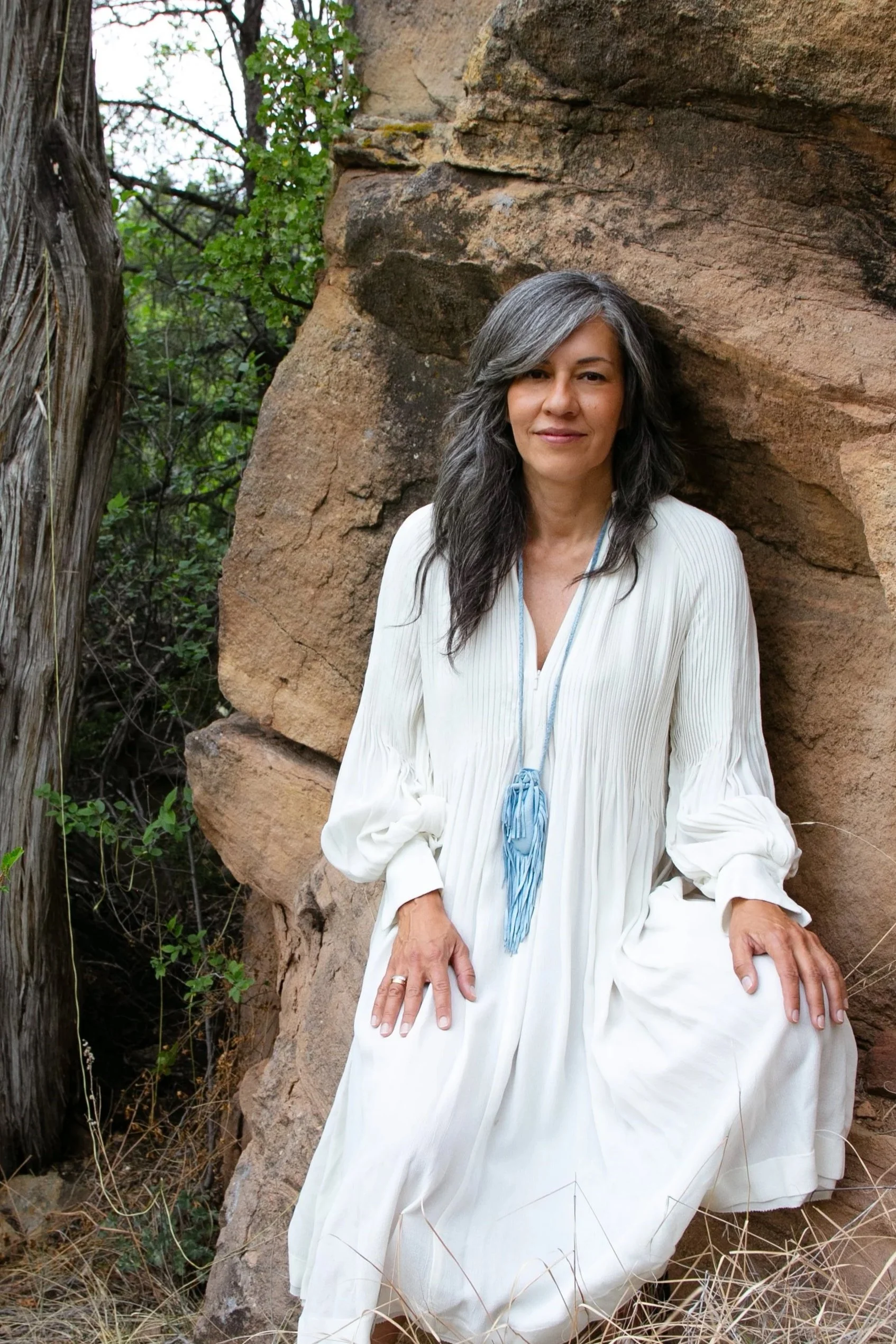 A woman with long gray hair and light skin sitting against a large rock outdoors, wearing a flowing white dress with her hands resting on her knees, surrounded by dry grass and green foliage.