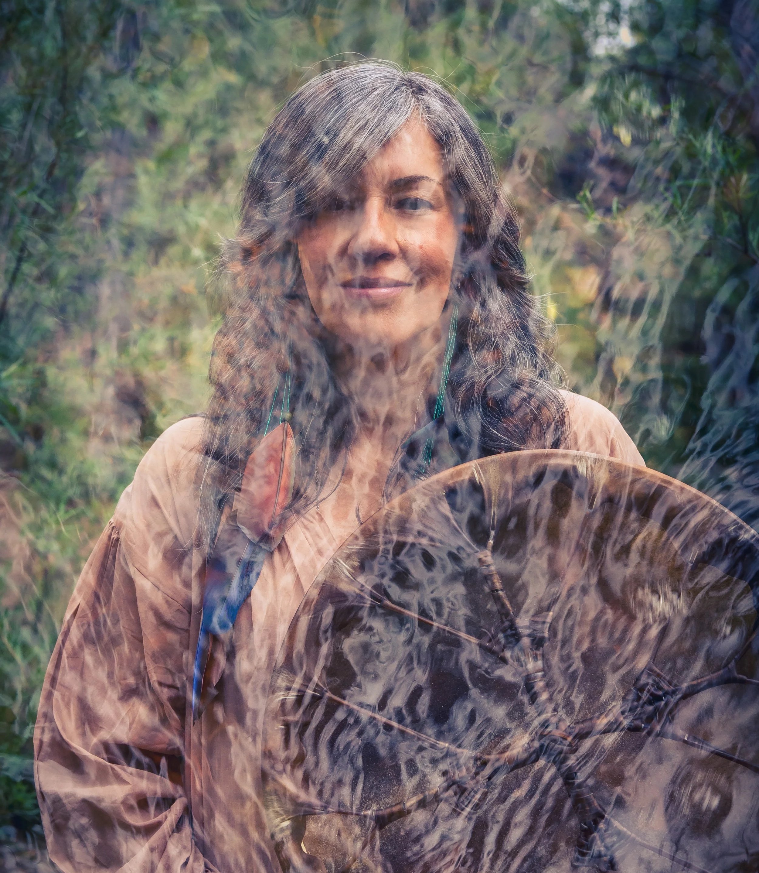 A woman with long gray hair smiling outdoors with shamanic drum and greenery in the background.