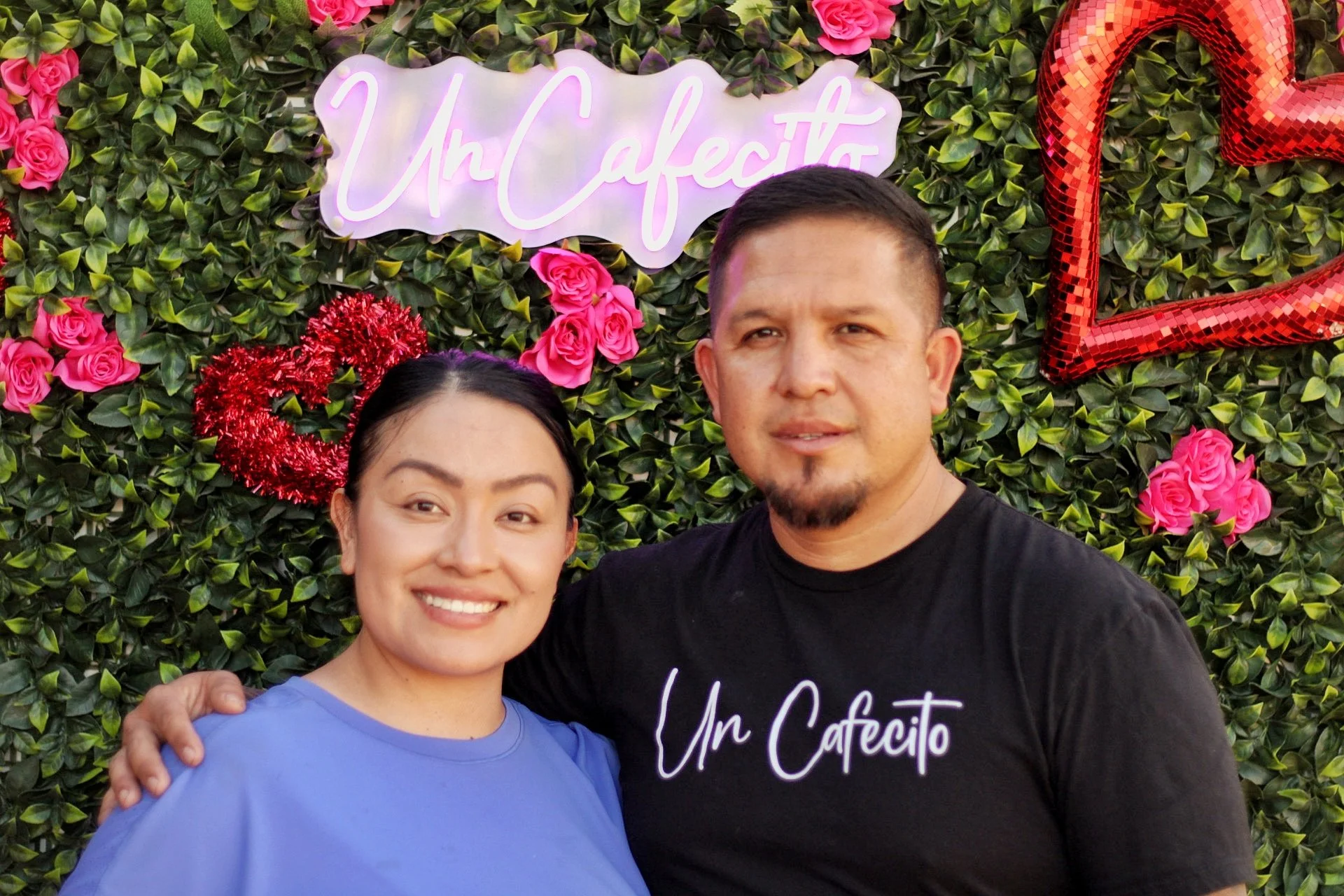 A smiling woman and a man taking a selfie in front of a decorative green wall with pink roses, a neon sign that says 'Un Cafecito,' and red heart-shaped balloons.