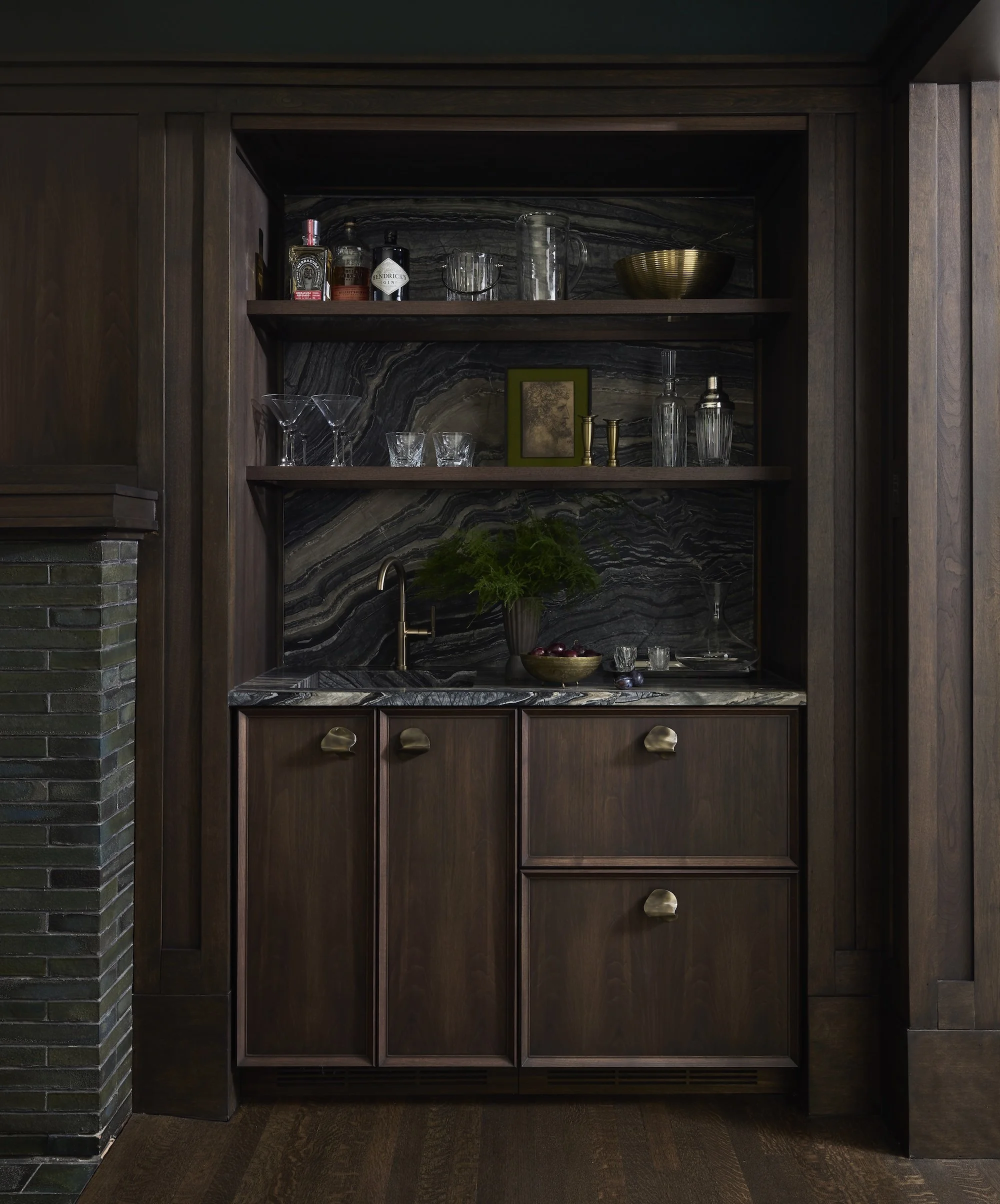 Dark wood bar cabinet with a marble backsplash, open shelves displaying various glassware and decorative items, a small framed picture, and a bowl of cherries on the lower shelf.