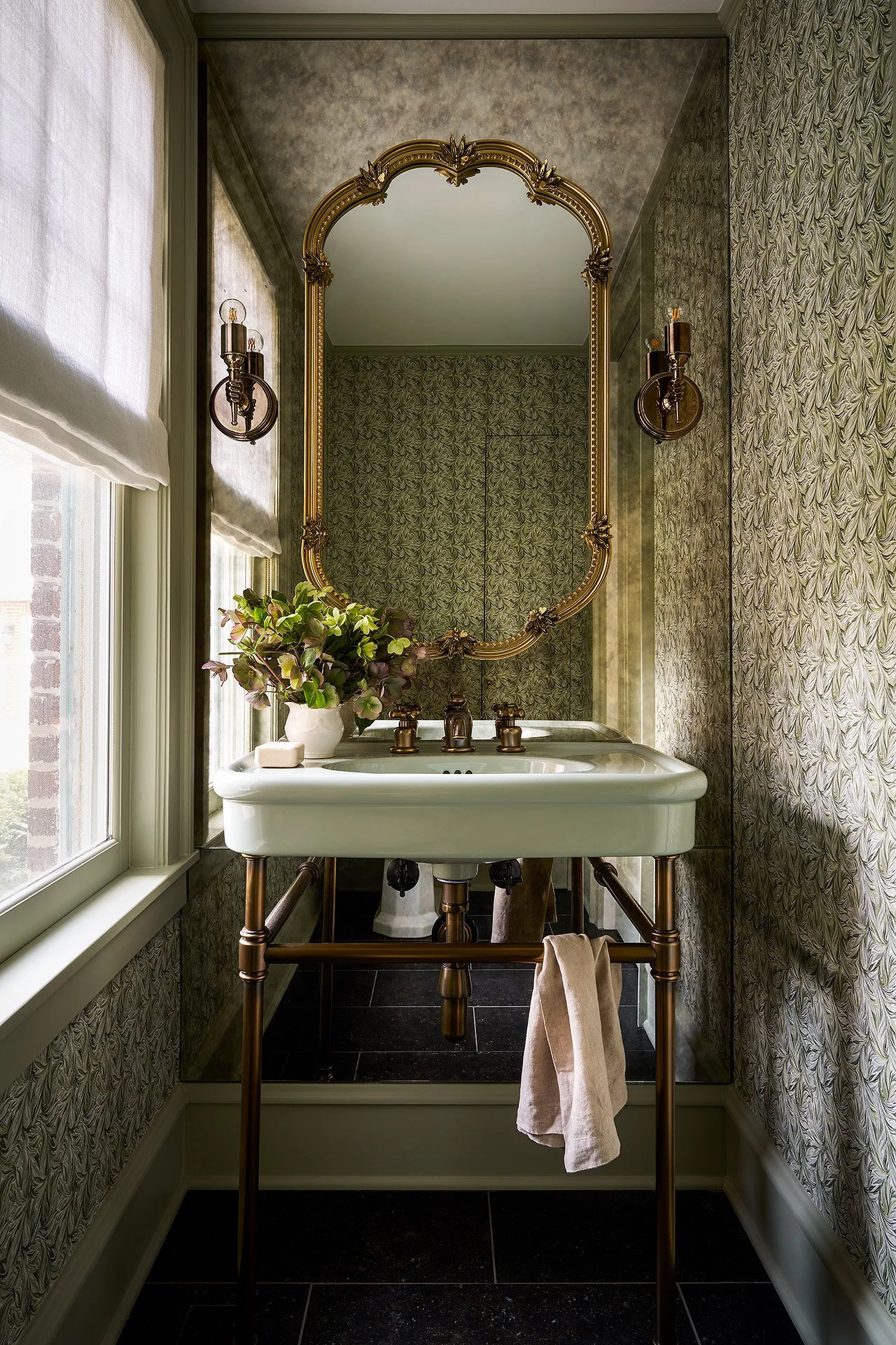 A vintage-style bathroom vanity with a white sink and gold fixtures, set under a large ornate gold-framed mirror, flanked by wall sconces, with patterned wallpaper, a potted plant, and a window with white blinds.