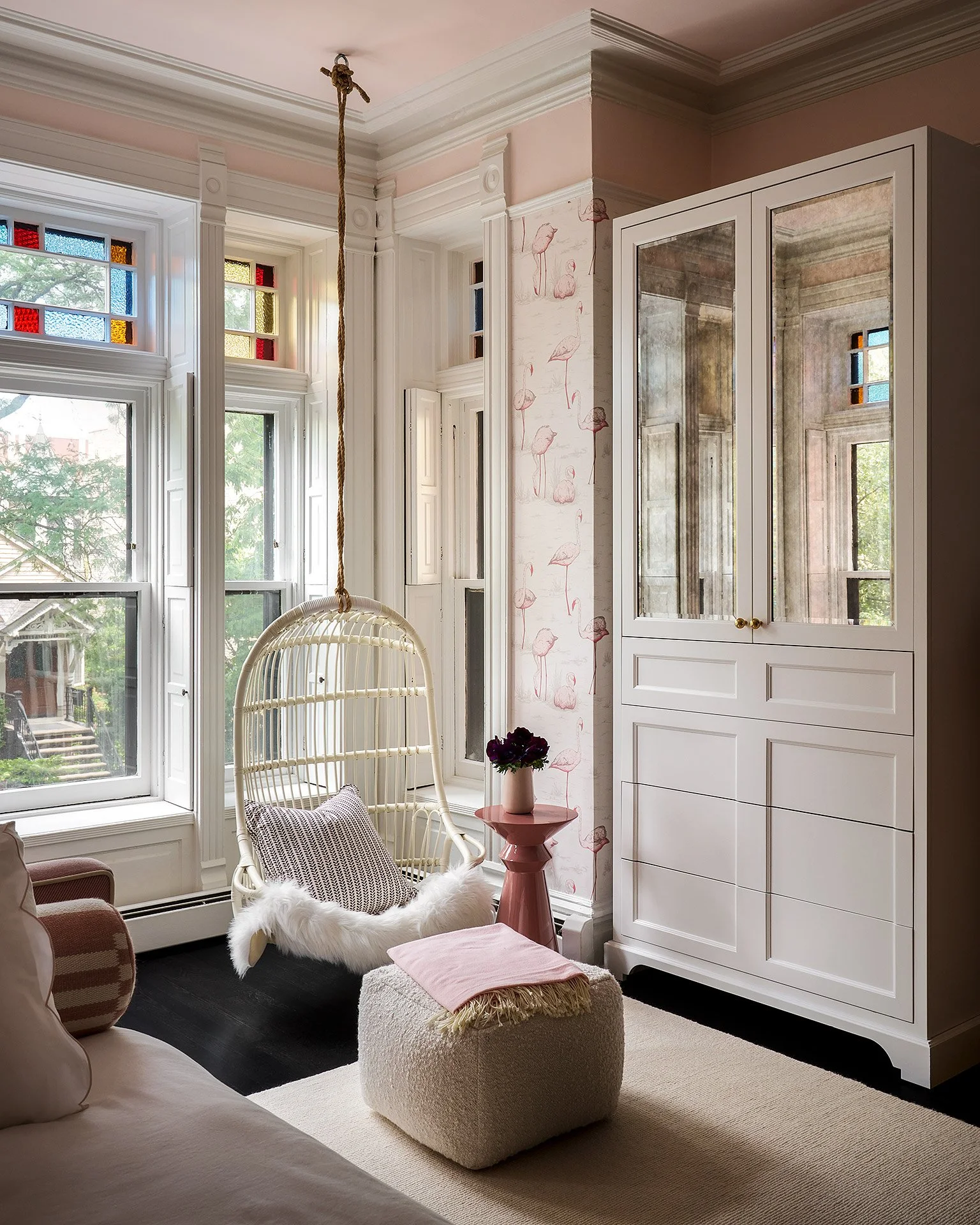 Sunlit living room with large windows, white cabinetry, a white hanging chair, a pink side table with a flower arrangement, a fuzzy ottoman with a blanket, and patterned and solid-colored pillows.