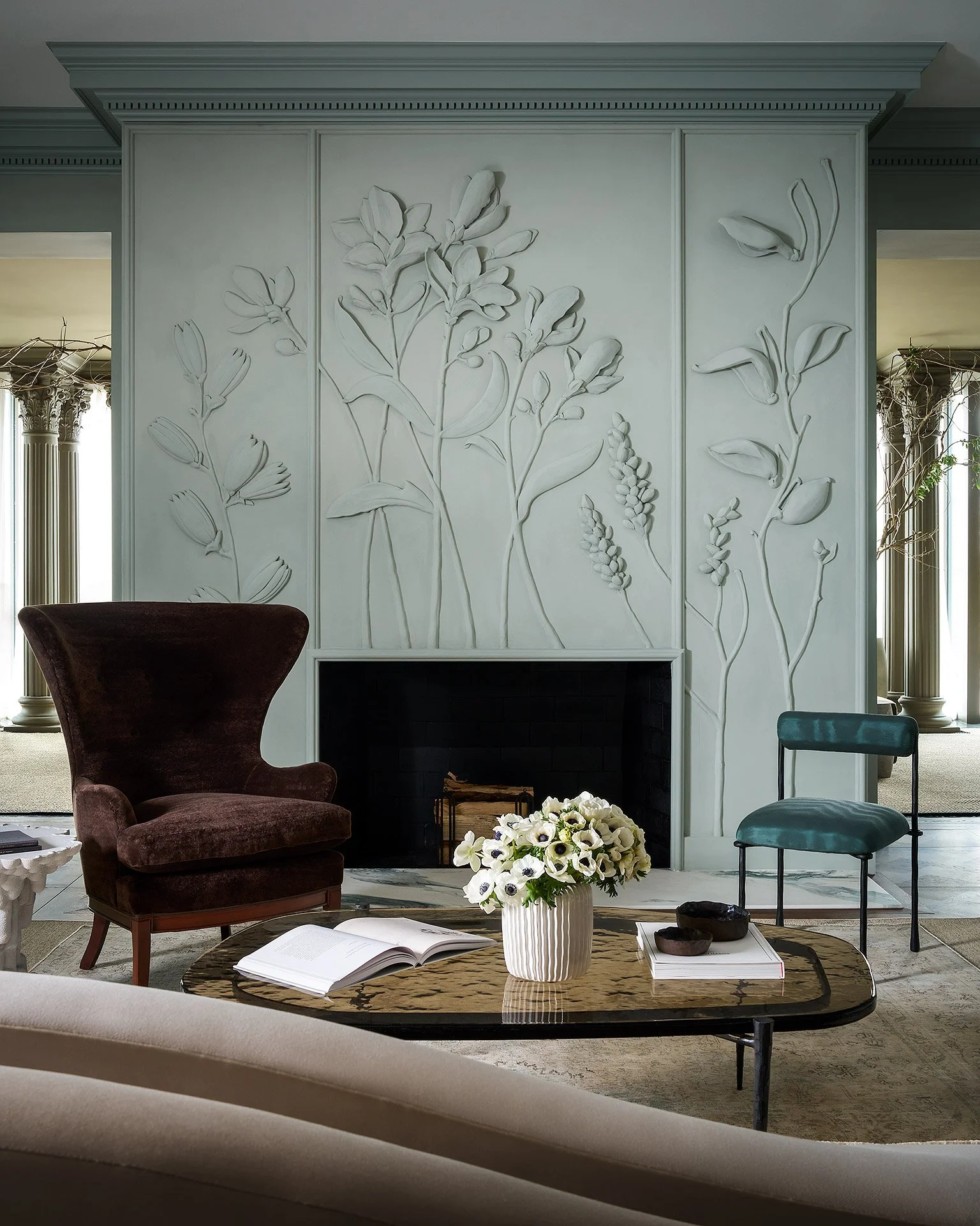 Living room with a white fireplace wall featuring floral relief design, dark brown armchair, blue-green chair, white vase with flowers, open book, and decorative bowls on a coffee table.
